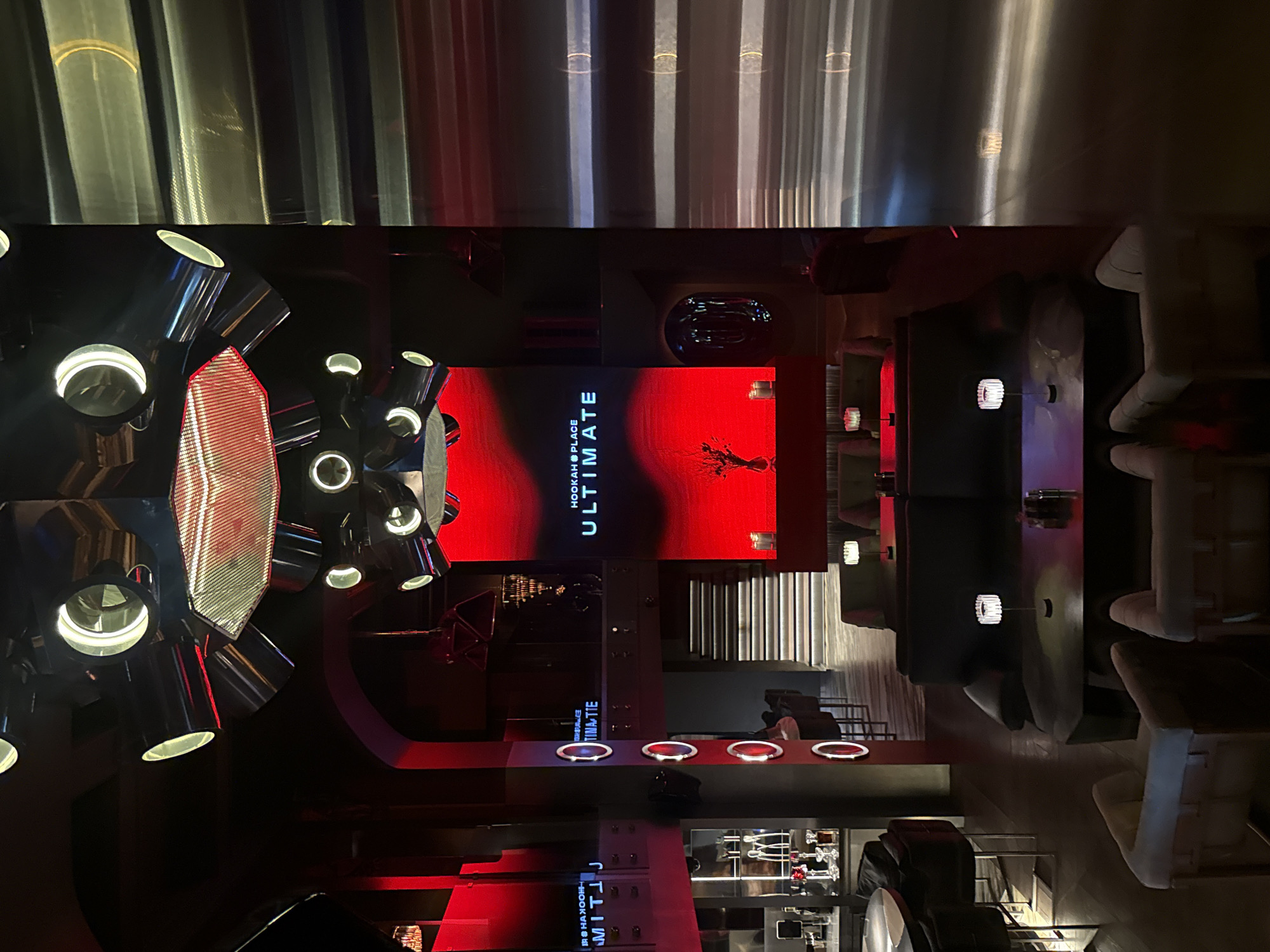 This image captures the interior of a modern, upscale lounge or club, likely a hookah bar, at night. The scene is dominated by a dark aesthetic with striking red and white accent lighting, creating a sophisticated and contemporary atmosphere.
At the center, a large, vertical LED screen prominently displays "HOOKAH PLACE ULTIMATE" in white sans-serif text against a vibrant red background. To the right of the text, a stylized, dark, abstract figure, resembling a skeletal creature or insect, is visible on the red screen.
To the left, a large, complex light fixture, possibly a chandelier, features multiple arms with cylindrical spotlights emitting bright white light. Its central section is an illuminated, textured panel. Below this, a long, dark counter or bar-like structure extends horizontally, embedded with several circular, glowing white and red lights. Further left, a display shelf showcasing various glass hookahs and accessories confirms the venue's nature.
On the right side of the image, dark, upholstered seating, including armchairs and tables, is arranged in intimate settings, subtly lit by small wall-mounted lamps. Lighter-colored chairs or stools are also visible in this area. A modern staircase, possibly made of metal or concrete, can be seen ascending in the background to the right of the main screen.
The upper part of the interior features vertical, reflective panels or curtains that create a dynamic striped pattern from diffused light. The overall impression is one of a chic, modern establishment designed for evening entertainment and relaxation. No individuals are present in the image. The location is Moscow, Russia.