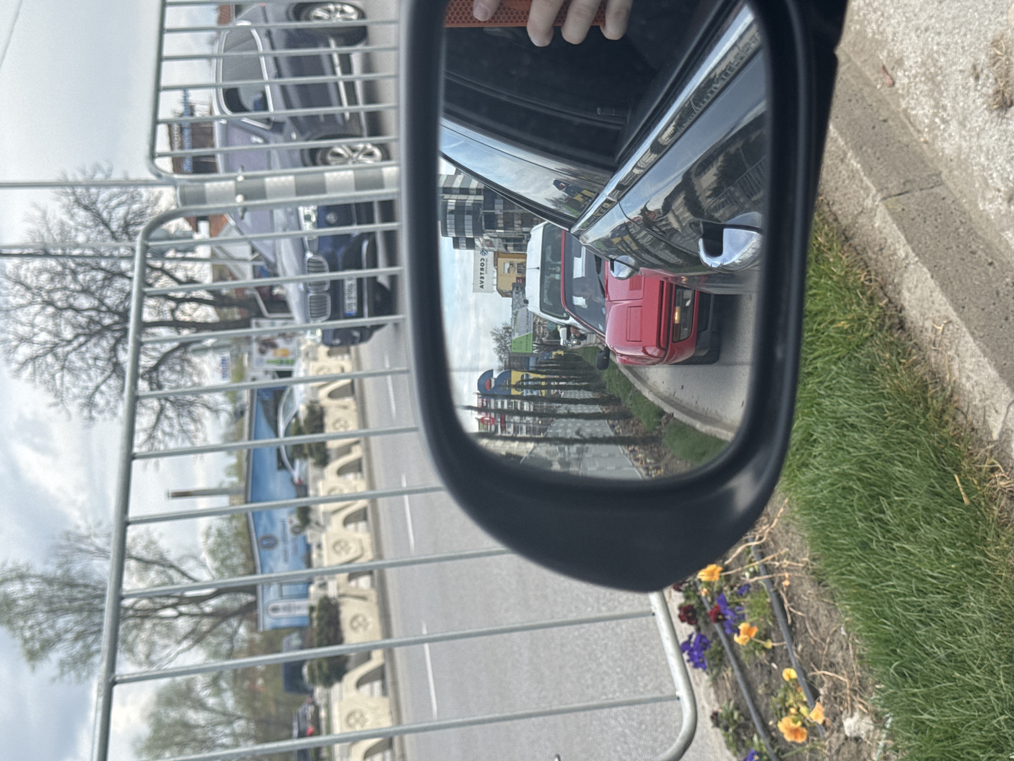 The image depicts a view from a car's side mirror, reflecting the road behind and adjacent to the vehicle. In the mirror's reflection, there are several cars, including a red car directly behind. There is also a white van in the reflection. A building with the word "СОБСТВЕНИК" and "COMEOV" can be seen in the mirror. Outside the car, a metal fence runs along the road. There is a curb with a flowerbed next to the car. In the distance and behind the fence are more cars and buildings. The location is likely a street in Plovdiv, Bulgaria, based on the Cyrillic writing on the building. There appears to be a hand visible in the upper left of the image, possibly the person holding the camera.