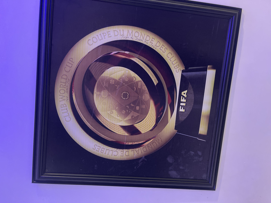 A framed picture depicting the FIFA Club World Cup trophy is mounted on a light-colored wall illuminated by a purple light source. The image, encased in a black frame, features a gold and dark-toned representation of the trophy. Inscriptions around the circular trophy design read "CLUB WORLD CUP," "MUNDIAL DE CLUBES," and "COUPE DU MONDE DES CLUBS." The base of the trophy image prominently displays "FIFA." Faint smudges are visible on the left side of the print within the frame. No individuals or active interactions are present. This scene is located in Orie Uratta, Nigeria.