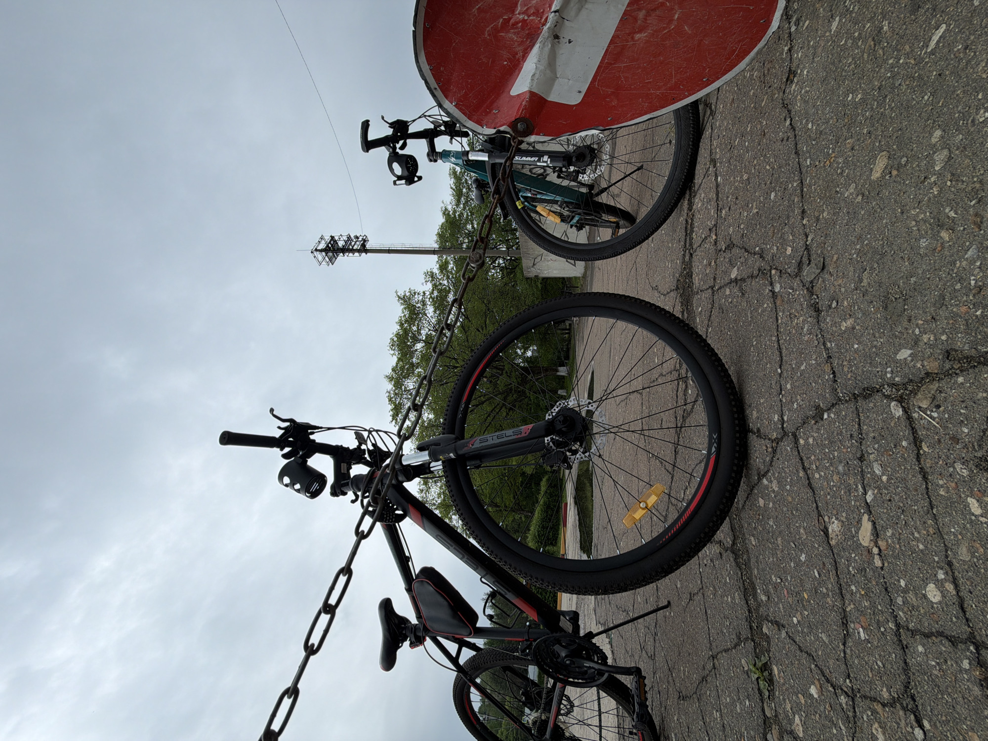 The image depicts two bicycles locked together with a heavy chain to a large, round, red and white "no entry" sign. The bikes are situated on cracked, grey pavement. In the background, a tall communications tower rises beyond the trees. Based on geolocation information, this scene is located in Birobidzhan, Russia. The primary subject is the security measure taken to prevent bicycle theft. No people are visible in the image.