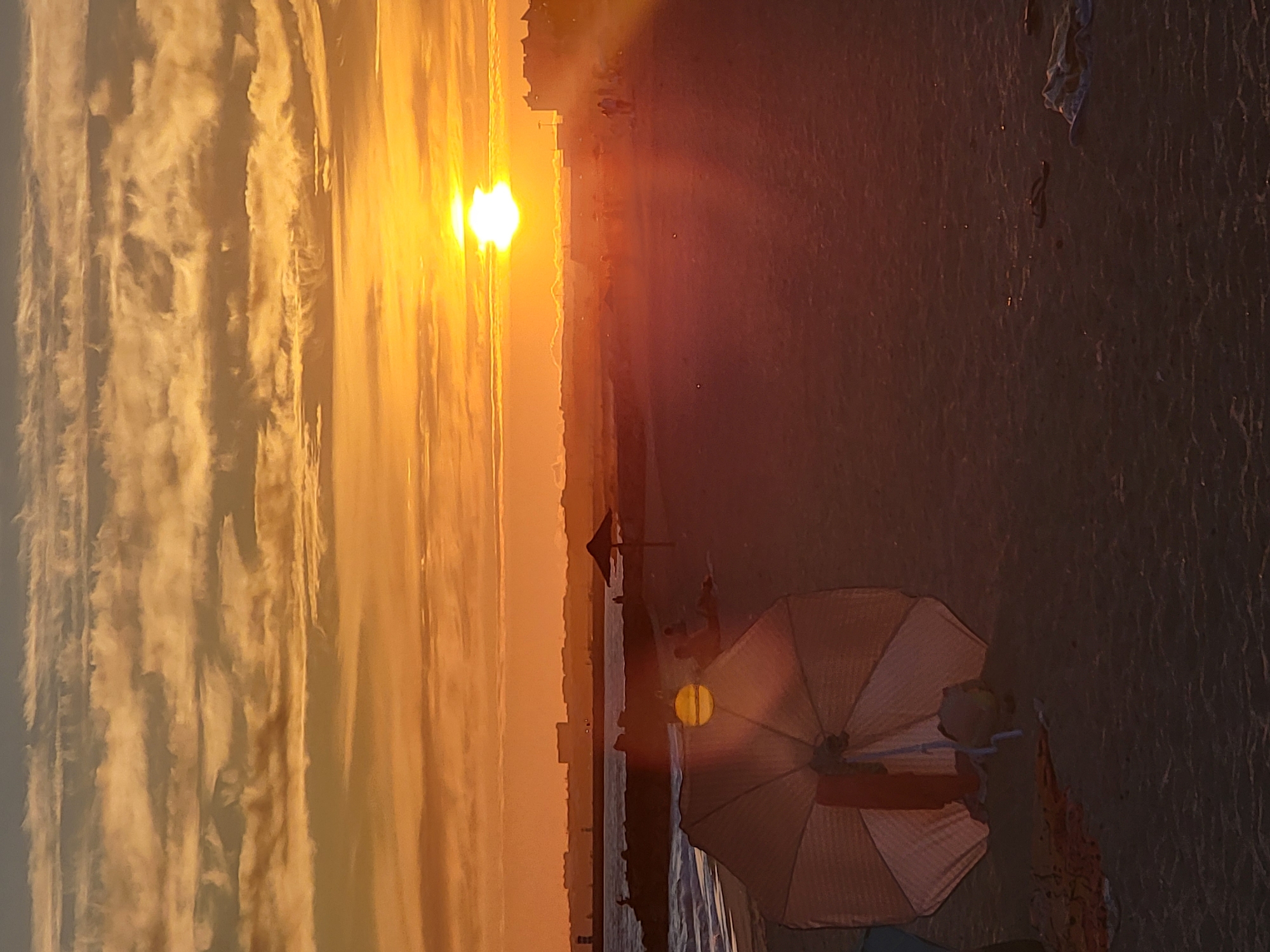 The image depicts a sunset scene on the beach in Quarteira, Portugal. The sun, a bright orb, is low on the horizon, casting a warm orange glow across the sky, which is filled with striated clouds. The foreground is dominated by a partially opened beach umbrella, its white fabric reflecting the sunset light. Nearby, a red towel and some other beach items are visible on the sand. In the mid-ground, several people are scattered across the beach, some seated, others walking. Two beach umbrellas are visible in the distance, along with a breakwater separating the sandy shore from the sea. The sea itself is calm, with gentle waves lapping the shore. In the background, the Quarteira skyline is silhouetted against the vibrant sky, featuring buildings and a lighthouse. The overall atmosphere is serene and tranquil, characteristic of an evening on the Portuguese coast.