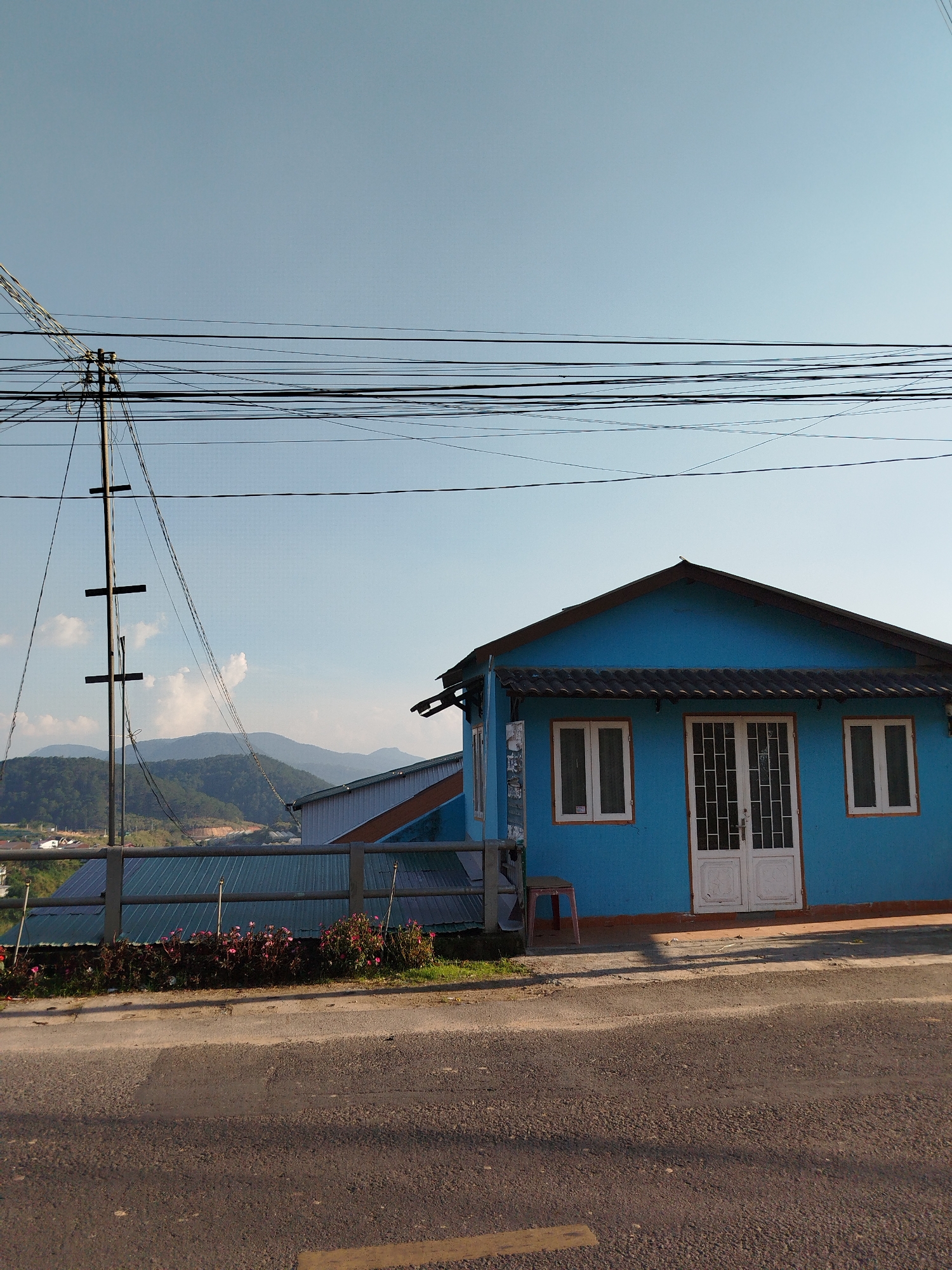 The image depicts a blue, single-story house with white trim and a dark roof in Da Lat, Vietnam. The house has a double door flanked by two windows. A small, reddish-brown stool sits beside the house. A fence and flower bed run along the front of the property. Behind the house, corrugated metal roofs of other buildings are visible, along with hills in the distance. A telephone pole with multiple wires stretches across the sky to the left of the house. The foreground is a paved road.