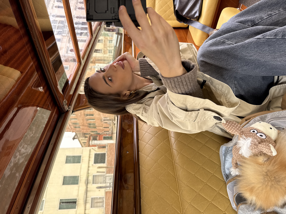 A young adult female is depicted inside a water taxi or private boat on a canal in Venice, Italy. She is positioned on the left side of the frame, holding a black smartphone with her right hand, seemingly taking a selfie or recording a video. Her left hand rests under her chin, and she looks towards the phone with a composed expression. She has long dark hair, is wearing gold hoop earrings, a light beige trench coat over a grey ribbed sweater, and blue denim jeans. The boat interior features polished dark wood trim and quilted mustard-yellow leather seating. To her left, a light grey pet carrier contains a small, light brown, plush horse-like toy. To her right, a dark shoulder bag rests on the seat. Through the boat's windows, classic Venetian architecture is visible, consisting of multi-story buildings with various window styles, some in light stucco and others with reddish-brown facades, implying a canal setting.
