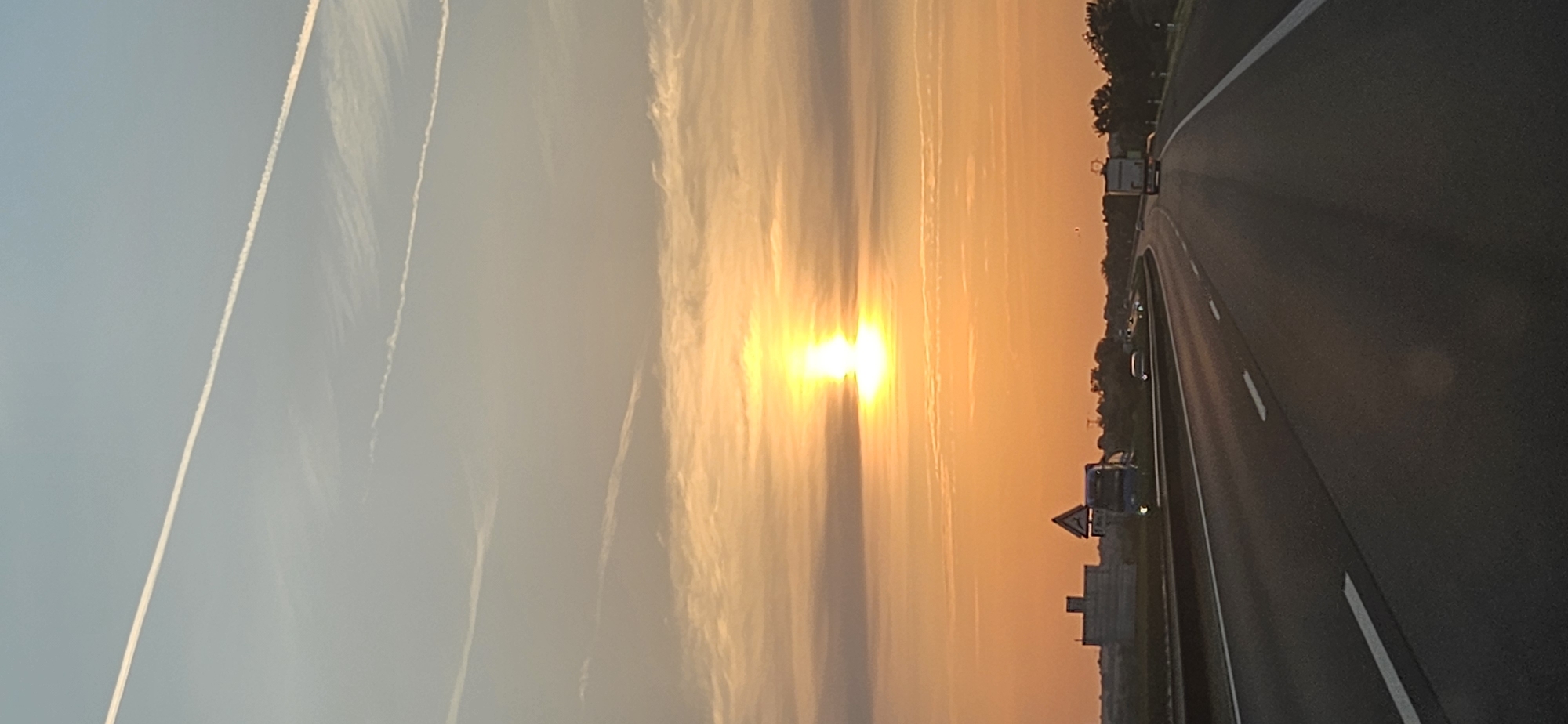 The image depicts a sunset view from a highway likely in or near Balatonfőkajár, Hungary. The sun, a bright orb, is setting low in the sky, casting a warm orange glow and illuminating thin, elongated clouds. Several faint contrails from aircraft are visible in the pale blue sky above.
The highway itself is a multi-lane road with clear lane markings. In the foreground, the road surface is visible, with shadows cast by unseen objects. In the distance, several vehicles are present on the highway, including what appears to be a truck in the right lane. On the left side of the highway, there is a large, boxy structure that might be a building or industrial facility, and a triangular road sign. Trees and vegetation line the sides of the highway, forming a natural backdrop to the scene. The overall impression is one of a tranquil evening drive.