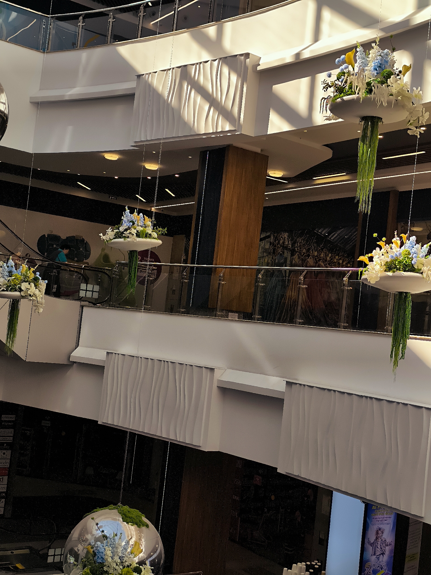 An interior shot of a multi-level shopping mall shows decorative floral arrangements hanging from the ceiling. The mall is brightly lit with natural light streaming in from above, casting shadows on the white walls and railings. In the foreground, a large, clear spherical ornament contains a floral arrangement of white and blue flowers with greenery. In the midground, two similar floral arrangements hang from above, one to the left and one to the right, each in a white basin with green hanging strands. A glass railing separates the levels, and a mural or large artwork is visible on the wall in the background. Escalators and people are also present on different levels of the mall. The image is taken from a high vantage point, looking down into the mall.