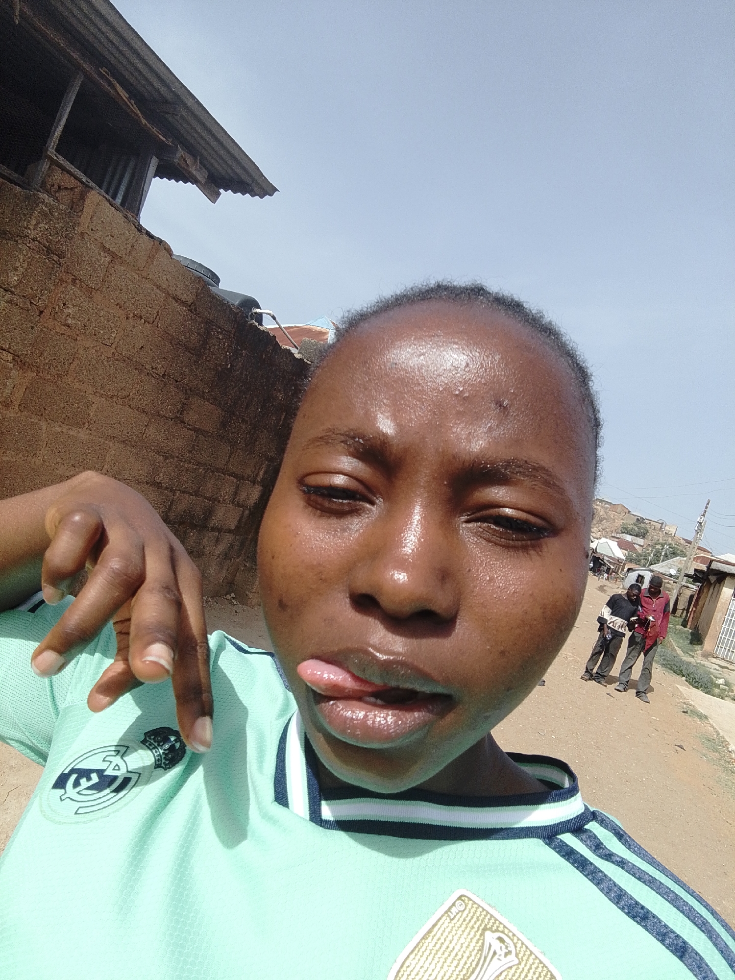 A close-up selfie captures a young person, likely female, making a playful expression by winking with their left eye and sticking their tongue out. Their skin is dark-toned and appears shiny, possibly from sweat, reflecting the bright sunlight. They are wearing a light mint green sports jersey with dark blue and white striped trim on the collar and shoulders. A visible logo on the left chest is identifiable as the "Real Madrid" crest, while another golden-colored crest, possibly a FIFA World Cup badge, is seen lower on the shirt. The person's right hand is raised near their chin, with fingers splayed in a gesture, possibly a peace sign or similar.
The background on the left features a weathered reddish-brown brick wall, topped by what appears to be a black water tank and part of a dilapidated corrugated metal roof structure. The right side of the background reveals an unpaved, dusty road stretching into the distance. Two adult males are visible standing further down this road. Beyond them, there are basic buildings or shacks, a utility pole, and distant hills under a clear, pale blue sky. The scene is brightly lit by the sun, indicating daytime. The environment suggests an outdoor, possibly residential or semi-urban setting, consistent with a location like Jos, Nigeria.