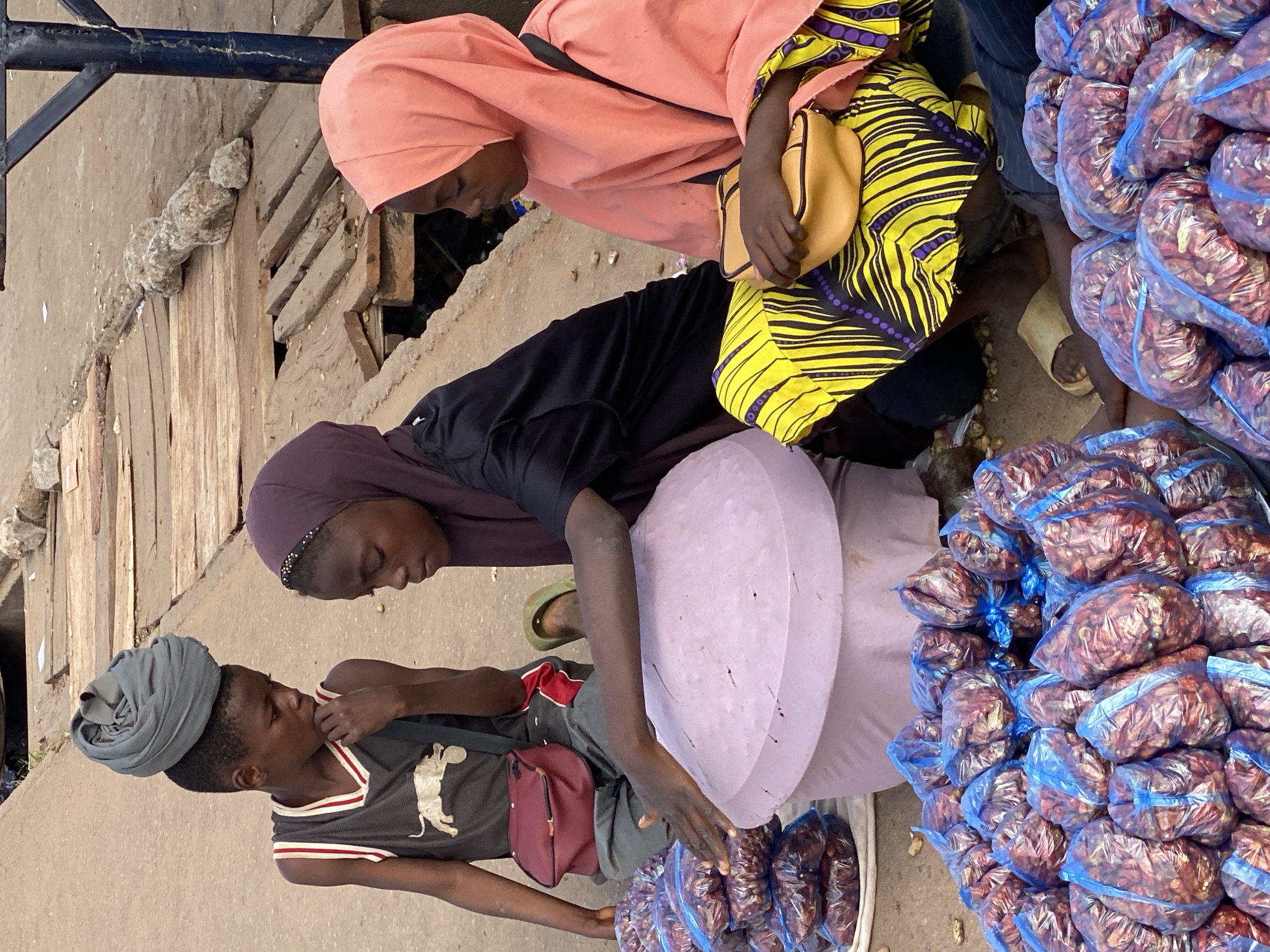 In Sabon Tasha, Nigeria, a market scene unfolds with three individuals interacting amidst a backdrop of goods. A person wearing a peach-colored hijab and a yellow and black patterned garment is seated, holding a small tan purse. Beside them, another person, also wearing a dark hijab, leans forward, their head down. Facing them is a young person with a gray turban on their head, dressed in a dark t-shirt with a white graphic and red trim. They are reaching out with their right hand towards a pile of items in plastic bags.
The objects surrounding the people are primarily produce. Large quantities of what appear to be dried chili peppers are bundled in clear plastic bags with blue ties, stacked high. Between the seated figures and the young person is a large, light purple, circular object, possibly a tray or a lid, also surrounded by bags of produce. The ground appears to be a mixture of concrete and dirt, with some wooden planks visible in the background.
The overall impression is one of a bustling market transaction or exchange. The lighting suggests it is daytime, likely in the morning or afternoon. The attire of the individuals, the style of the market stall, and the produce point to a West African setting. The focus of the interaction seems to be on the sale or purchase of the goods. There is no visible text in the image.