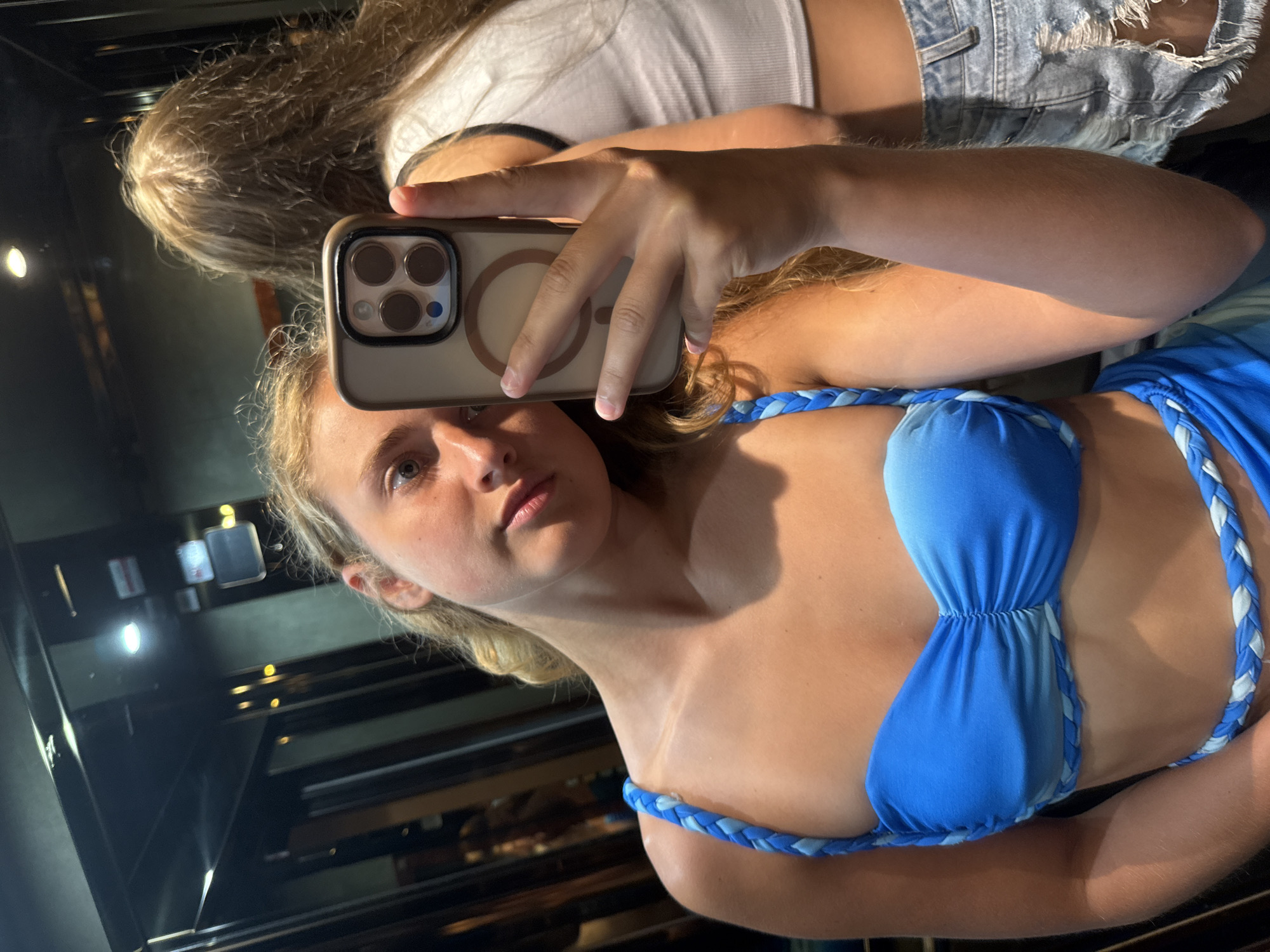 The image shows a young woman taking a selfie in what appears to be an elevator with mirrored walls. She is wearing a blue bikini with braided straps and a matching sarong or skirt. Her blonde hair is down and wavy. In the reflection of the mirror, another young woman with blonde hair is visible, wearing a white tank top and ripped denim shorts. The elevator is dimly lit, with some lights visible on the ceiling and walls. The overall impression is of a casual, recreational setting, possibly on vacation. The specific location in Trieste, Italy, cannot be determined from the provided image.