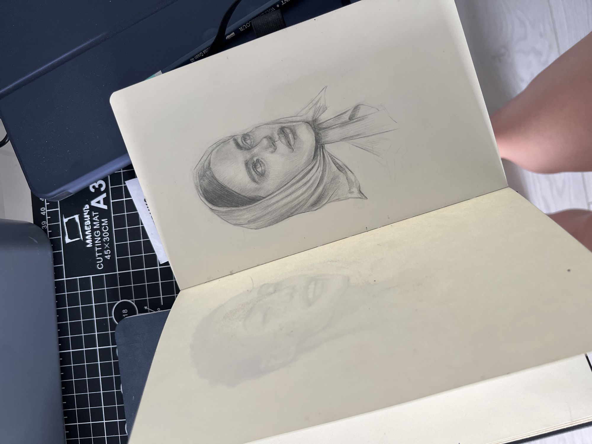 An open sketchbook is pictured, with a faint pencil sketch of a person's face on the left page. The right page features a more detailed pencil sketch of a woman wearing a headscarf, looking upwards and to the right. The sketchbook is placed on a black cutting mat with white grid lines. A dark blue tablet case is visible in the background, along with other office supplies. The overall scene appears to be indoors, with a person's legs visible at the bottom of the frame.