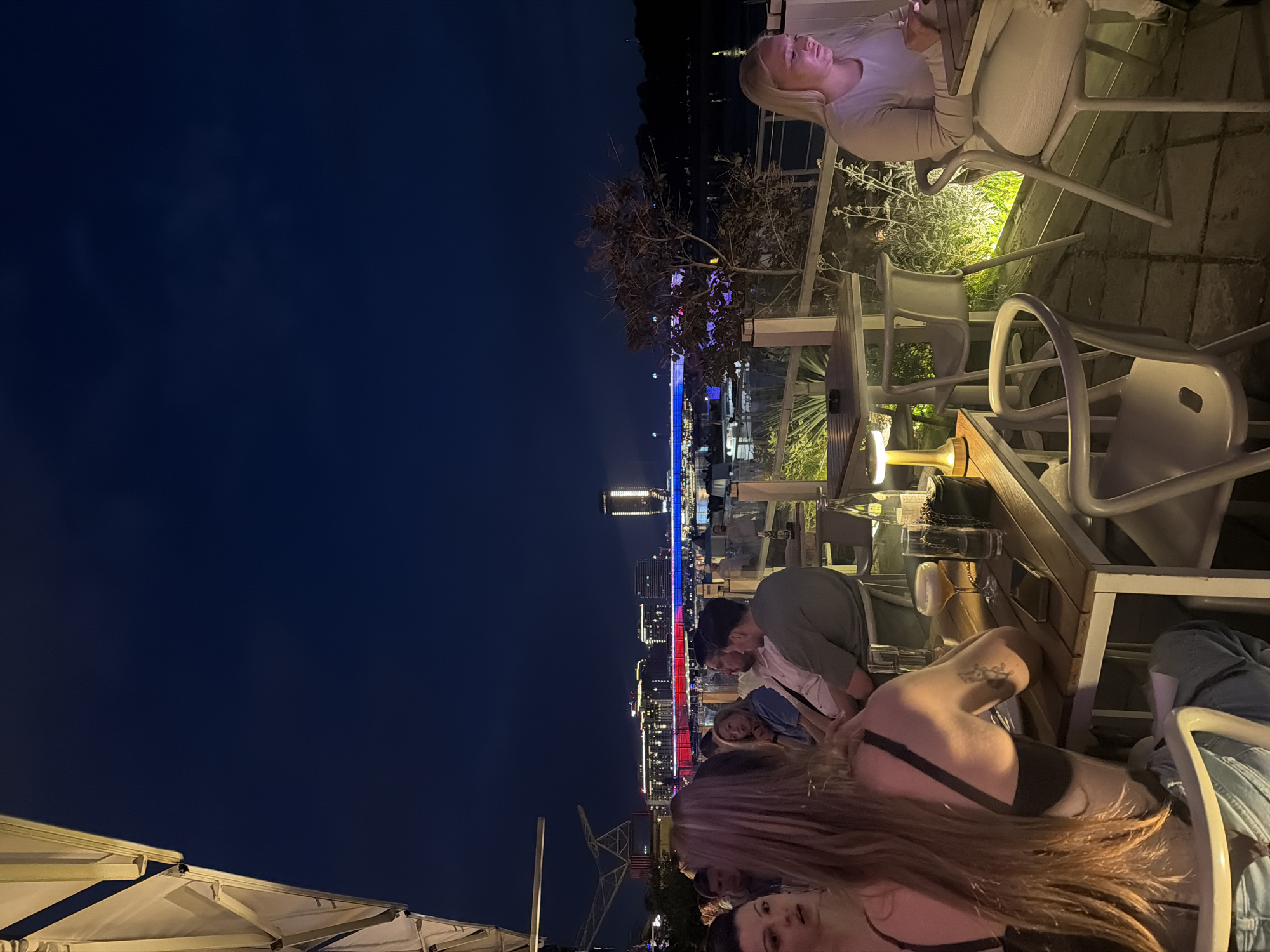 The scene shows an outdoor dining or social setting at night. People are seated at tables, with some engaged in conversation or using mobile devices. Visible in the background is a cityscape illuminated with red, white, and blue lights, which suggests Belgrade, Serbia, due to its frequent use of these colors on buildings and bridges. Tables and chairs are modern and mostly light in color. One person is seen on the top of the image using the phone.