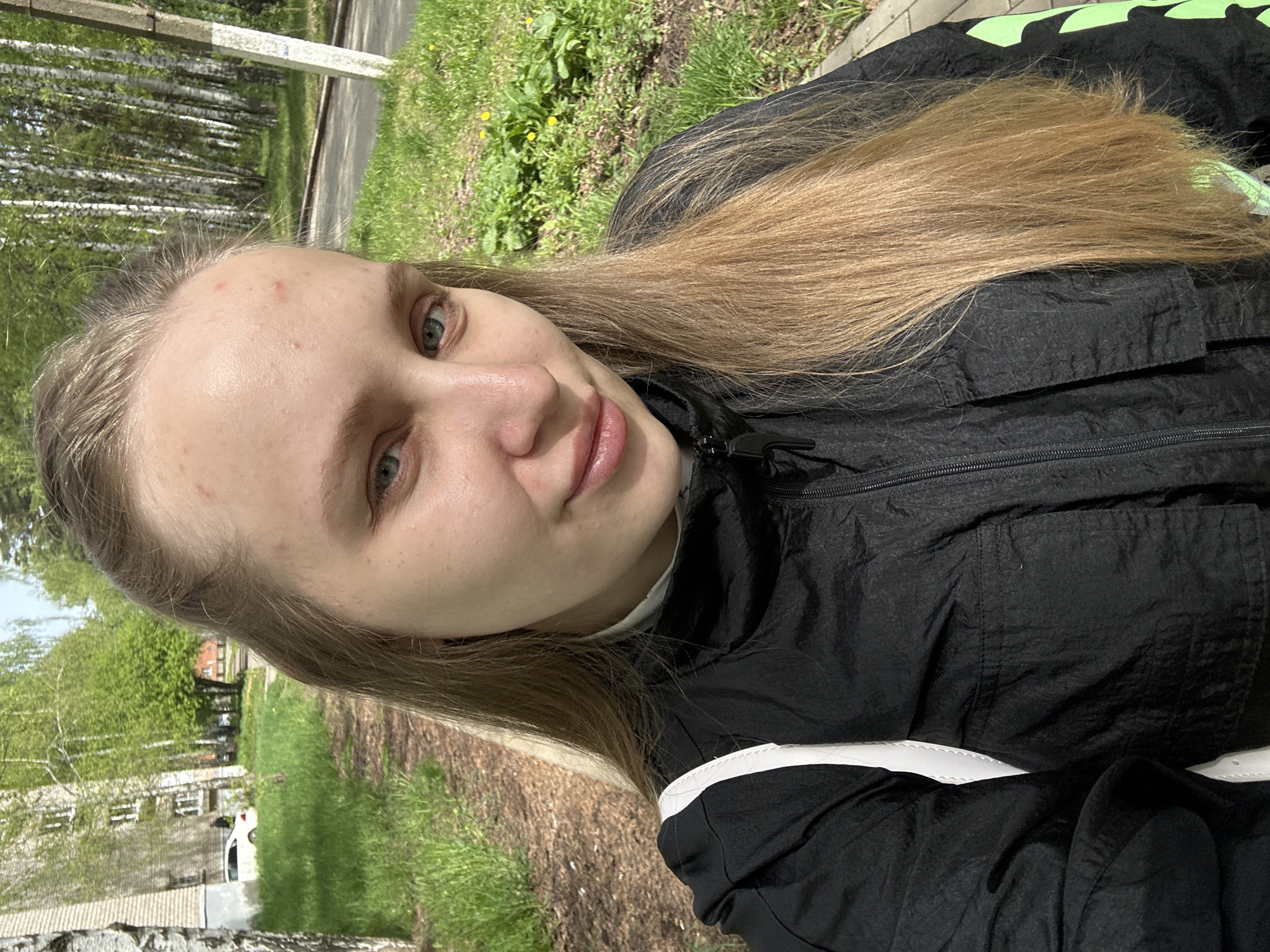 A young woman with blonde hair is posing for a selfie outdoors. She is wearing a black jacket with a white stripe along the shoulder and a white collar underneath. She has light skin with some minor blemishes on her forehead. She is gazing slightly towards the left. The background shows a park-like setting with trees and grass. The image was taken at Chaikovsky, Russia.