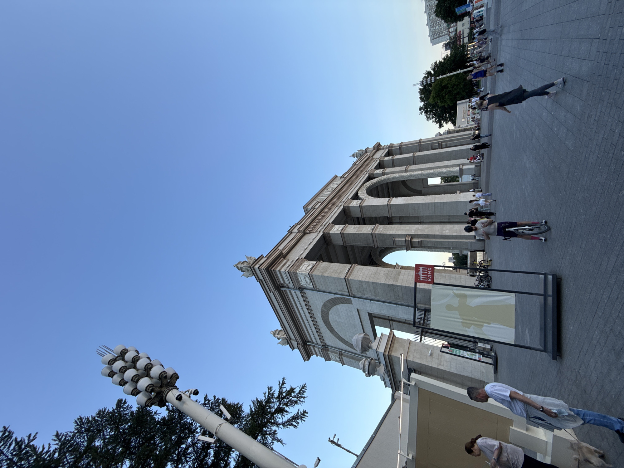 The image depicts the Main Entrance Arch of VDNKh (Exhibition of Achievements of National Economy) in Moscow, Russia. The grand archway, constructed from light-colored stone with classical architectural elements, dominates the right side of the frame. Its multiple arches and columns are clearly visible against a clear blue sky. To the left, a distinctive lamp post with a cluster of circular lights rises high, partially obscuring a coniferous tree.
In the foreground, a paved plaza area is populated with several individuals. On the far left, a woman with dark hair is holding a drink and looking to her right. Beside her, a man in a white t-shirt and blue jeans stands leaning forward, holding a bottle and a rag. A small dog is visible near the man's feet. A display easel with a yellow silhouette on a white background stands in the center of the plaza. Further into the plaza, people are seen walking, with one person on a bicycle in the mid-ground. In the background, more people are milling about, and buildings can be seen beyond the trees and the arch. The lighting suggests it is late afternoon or early evening.