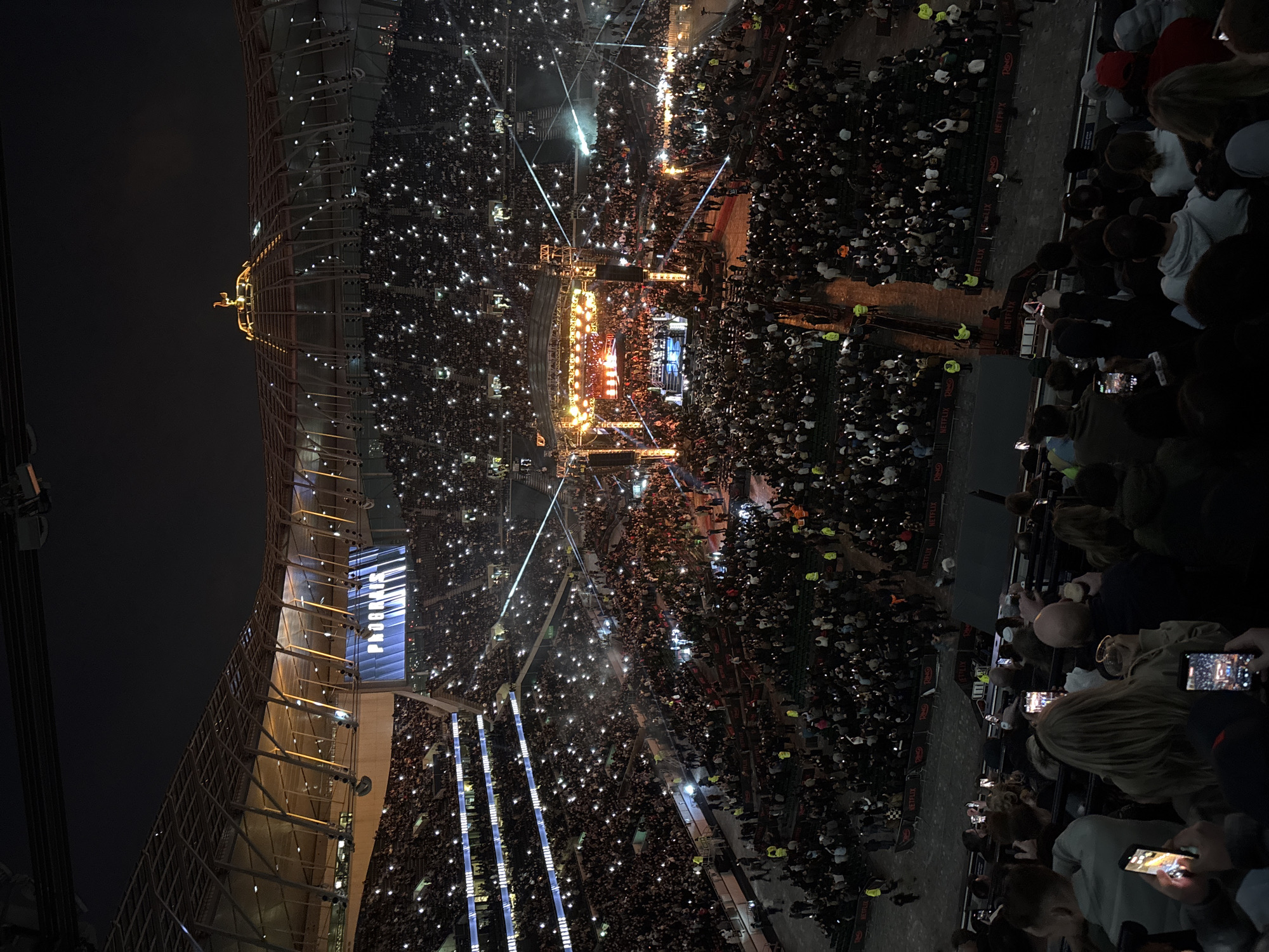 This aerial shot captures a large crowd at a stadium event at night. The crowd occupies the stadium's stands and floor, holding up illuminated devices that scatter light across the scene. A stage is set up in the center with visible lighting and what appears to be a performance in progress. The architecture of the stadium is partly visible, with the structure's exterior frame illuminated along the left side of the shot. Signage with logos of "Netflix" and "RING" are visible around the stadium. "PROGRAIS" is visible written on the side of the stadium, suggesting the location is the Tottenham Hotspur Stadium in London, United Kingdom. The darkness of the sky suggests it is nighttime.