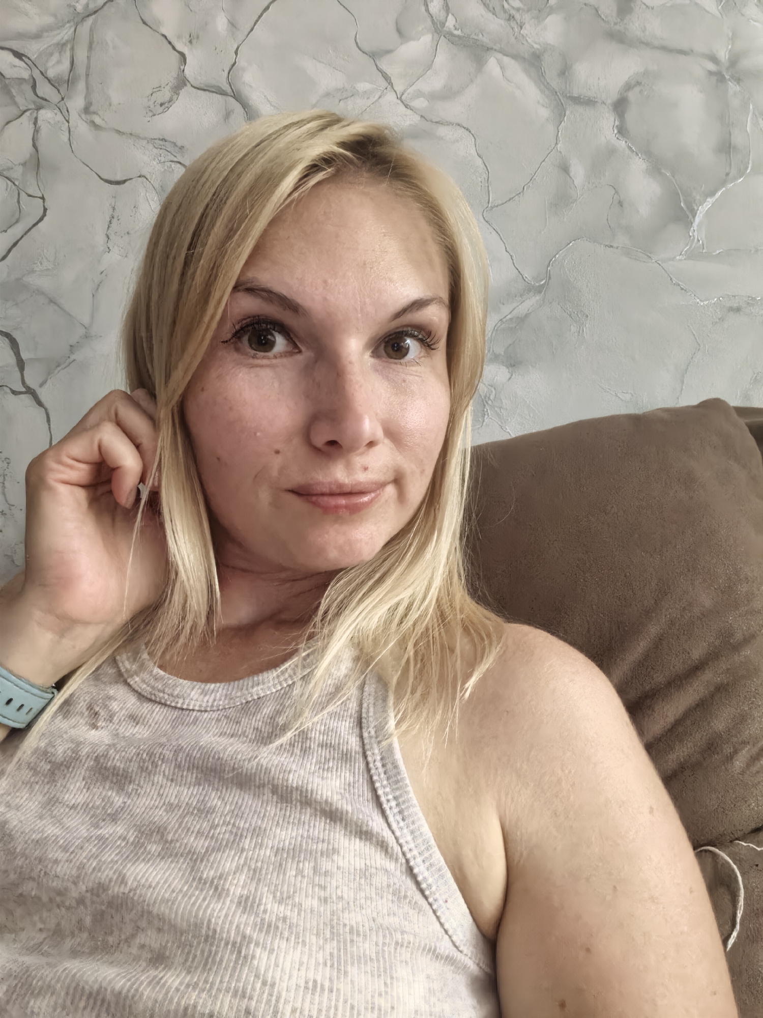 A woman with blonde hair and brown eyes is taking a selfie. She is wearing a gray tank top and a blue watch on her left wrist. She is sitting on a brown couch in front of a marble-patterned wall. Her right hand is touching her hair near her ear. The image is a medium shot, with the woman's face and upper body visible. The lighting is natural, and the background is slightly blurred. The location is not specified in Sterlitamak, Russia.