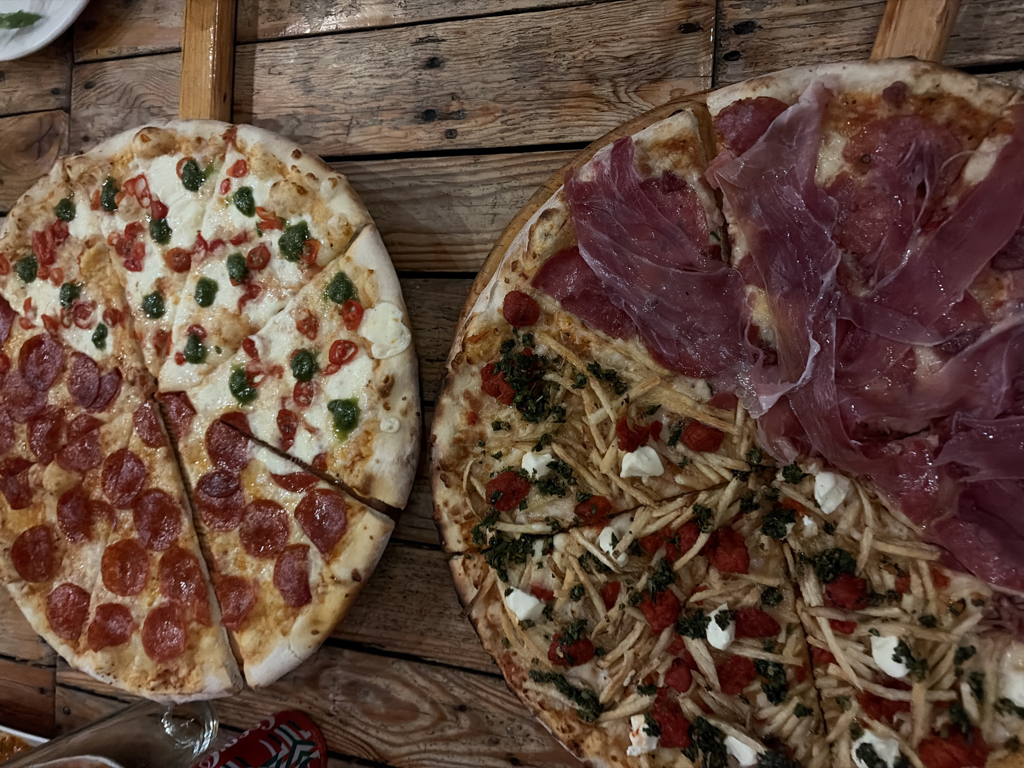 Here's a matter-of-fact description of the image:
The image shows two pizzas on a wooden table, photographed from an overhead perspective.
On the left is a round pizza, with half topped with pepperoni and the other half with mozzarella, red cherry tomatoes, and green pesto. Several slices have been cut.
On the right, a second pizza rests on a wooden board. Half of this pizza is topped with prosciutto. The remaining half is covered with long, thin fried potatoes, interspersed with red cherry tomatoes, green herbs, and dollops of white cheese. Several slices have been cut from this pizza as well.
The image does not provide information about the location of Unknown, Unknown.