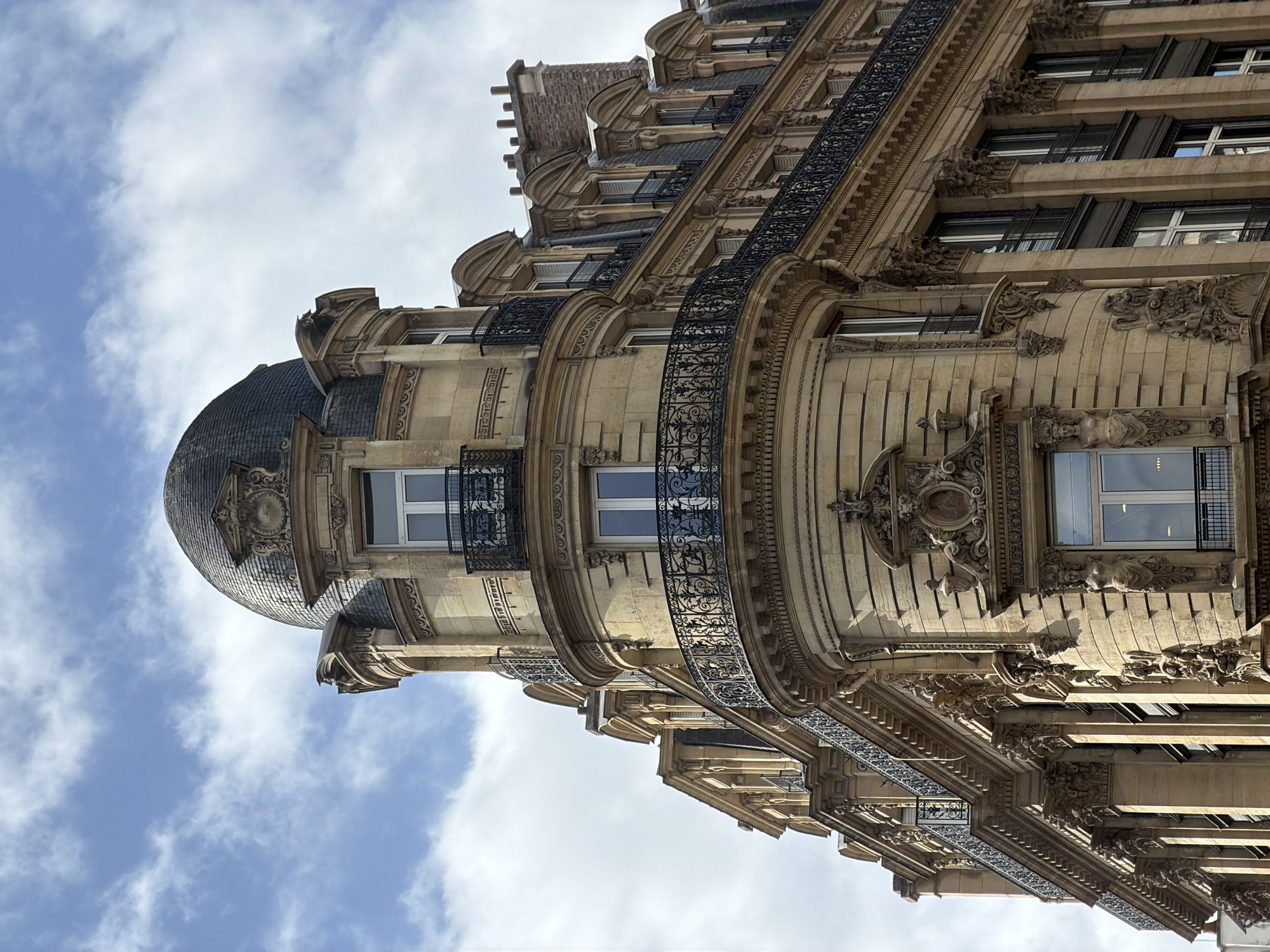 This is a low-angle shot of a classic Parisian building, likely dating to the late 19th or early 20th century, known for its ornate architecture and detailing. The building has a domed roof covered in dark, weathered tiles, with multiple windows visible on the turret-like structure and the adjacent facade. Black wrought-iron balconies with intricate designs accent the curved architecture. The facade is adorned with decorative carvings, including floral motifs and figurative sculptures beneath the window ledges. The building has a beige or light-brown stone exterior. The sky is visible in the background, with patches of light blue interspersed with puffy white clouds. The overall impression is one of Parisian architectural grandeur and detail.