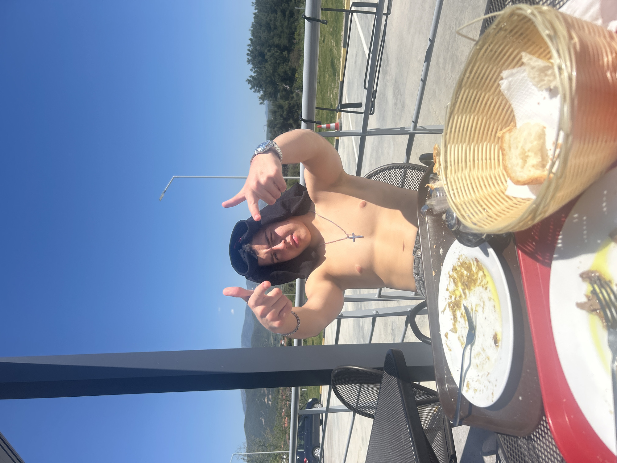 A young man is seated at an outdoor table in Νέα Κερδύλια, Greece, shirtless and wearing a black hat. He is making hand gestures, pointing his fingers up towards the camera. He wears a watch on his left wrist and a cross necklace. A plate with remnants of a meal and a bread basket are on the table in front of him. In the background, a building structure and surrounding landscape can be seen under a clear, blue sky.