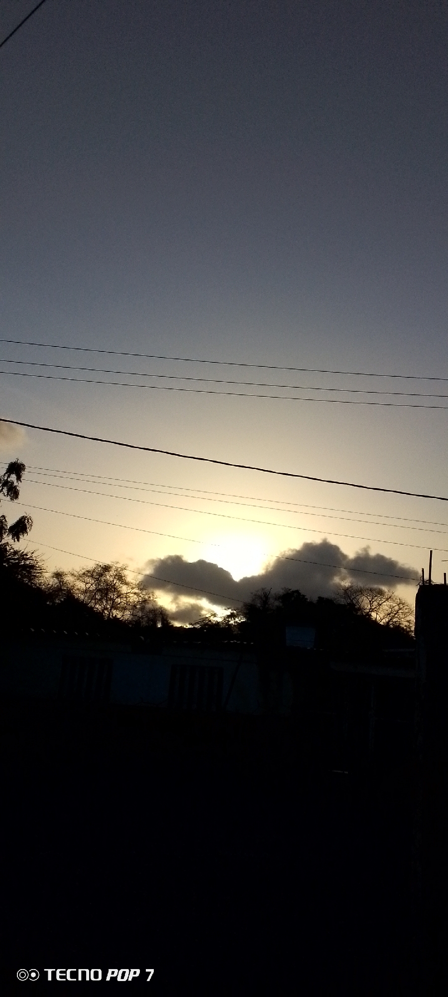 The image shows a sunset in Altagracia, Venezuela. The sky is a gradient of light gray to yellow where the sun is setting behind a layer of dark clouds. Silhouetted against the light are trees, a building, and power lines. The lower part of the image is in shadow, making the foreground details hard to distinguish. In the bottom left corner, the watermark "TECNO POP 7" is visible.