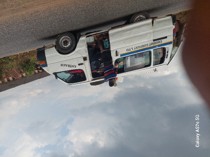 A white passenger van, identified by "ENTRACO" on the front door and "ENUGIRT COMPANY LTD" on its side, is stopped on an asphalt road in Awkuzu, Nigeria. The vehicle's passenger-side sliding door is open, revealing multiple occupants inside. One individual, wearing a striped garment with red, blue, and white bands over a dark shirt, stands outside the open door, facing into the van, indicative of boarding, alighting, or interaction with passengers. The van shows signs of operational wear. The environment consists of a dark asphalt road surface, a dirt shoulder with sparse green vegetation to the vehicle's right, and a slightly elevated background feature possibly incorporating a concrete or earth embankment with visible metallic elements. The sky above is partly cloudy.