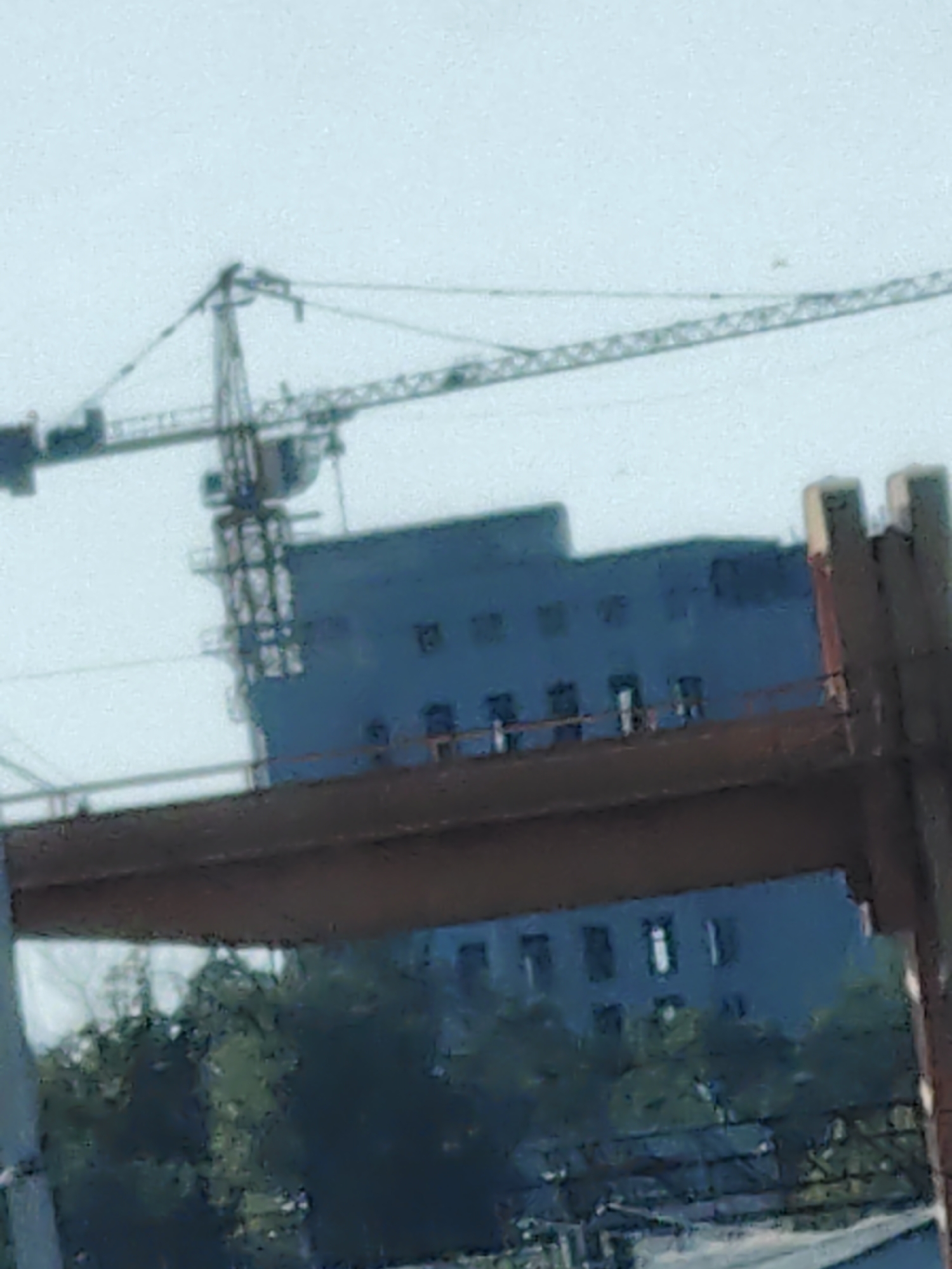 The image captures a construction site with a prominent crane towering over a dark blue building. The building appears to be a multi-story structure with visible windows. In the foreground, a thick, reddish-brown beam or bridge-like structure cuts across the frame. Below this structure, trees and foliage are visible, suggesting an outdoor environment. The sky is a hazy, light blue. The scene suggests ongoing construction or infrastructure development within the city of Shymkent, Kazakhstan.