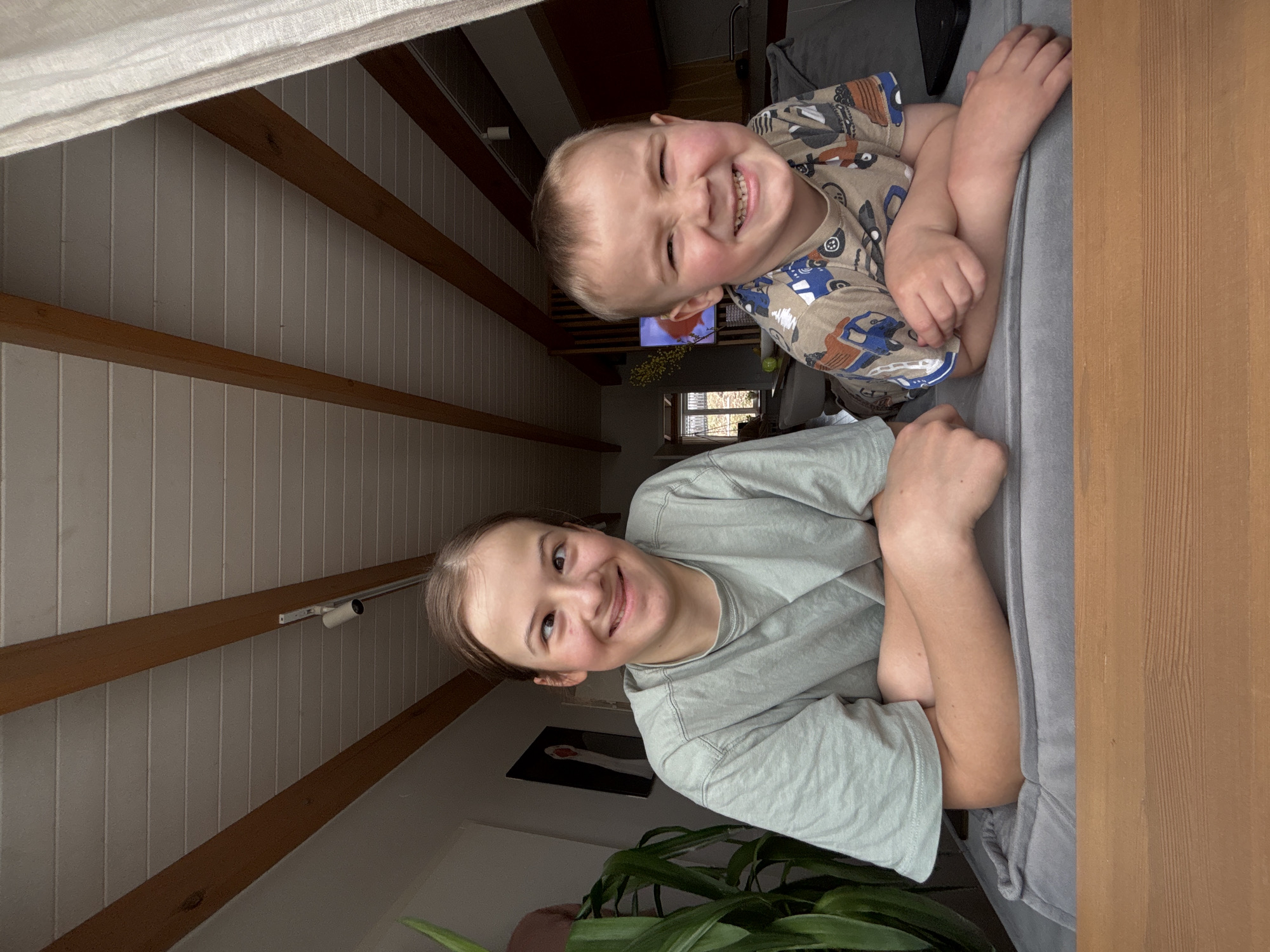 Two children, a girl and a younger boy, are pictured close together in a residential interior. The boy, located at the top of the frame, is fair-skinned with blond hair, and he's grinning, revealing his teeth. He wears a short-sleeved shirt with a pattern of vehicles. The girl, at the bottom of the frame, has light skin and brown hair pulled back. She is wearing a light green t-shirt and is smiling. Both are leaning against what appears to be a sofa or cushioned structure.
The background features a white paneled wall with exposed wooden beams, and a glimpse of a living area can be seen through an opening. A television is visible in the distance, along with some green plants and other furniture. To the left and behind the girl is a potted plant. The wall has a picture hanging on it. A white curtain is visible at the top of the frame. The vertical orientation of the image suggests it was taken from a mobile device.
There is no definitive visual information in the image to suggest it was taken in the city of Chaikovsky, Russia.