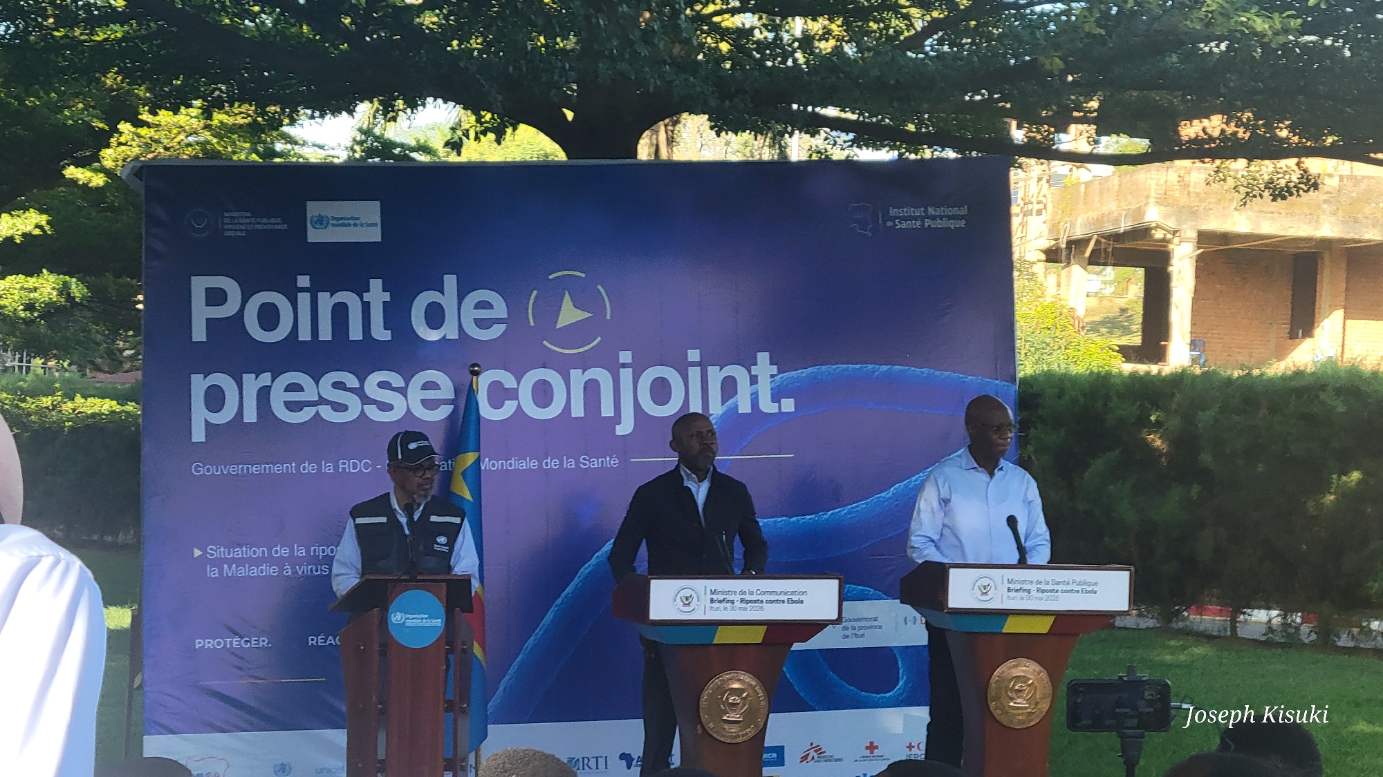 The image captures a joint press conference in Bunia, Democratic Republic of the Congo, likely related to public health concerns. Three men stand behind podiums, addressing an unseen audience. The backdrop is a large banner titled "Point de presse conjoint" (Joint Press Conference) with logos of the World Health Organization and the Institut National de Santé Publique (National Institute of Public Health). A flag of the Democratic Republic of the Congo is visible behind the man on the left. The podiums are branded with the ministry logos and a briefing title related to the Ebola response. A camera is positioned to record the event. Behind the banner, there is greenery and a partially constructed building. A watermark with the name "Joseph Kisuki" is visible at the bottom right.