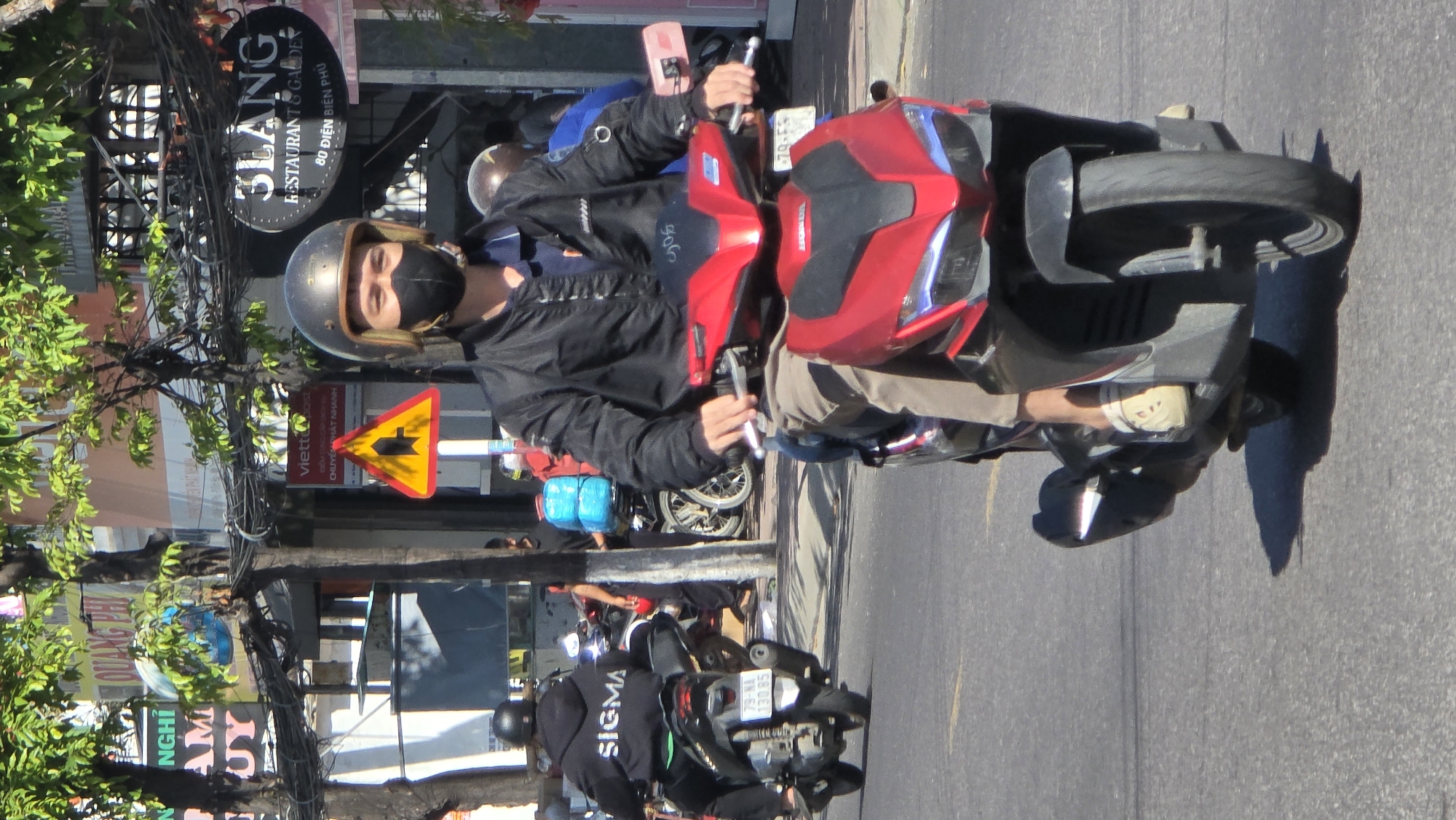 The image depicts a person riding a red Honda motorbike on a city street in Bắc Nha Trang, Vietnam. The rider wears a black jacket, khaki pants, white sneakers, a helmet, and a black face mask. Behind the rider is a sign for the restaurant "3Lang" at 80 Điện Biên Phủ street and a yellow traffic sign. Other motorbikes and people are visible on the street. The scene appears to be taken during the day with sunlight illuminating the street.