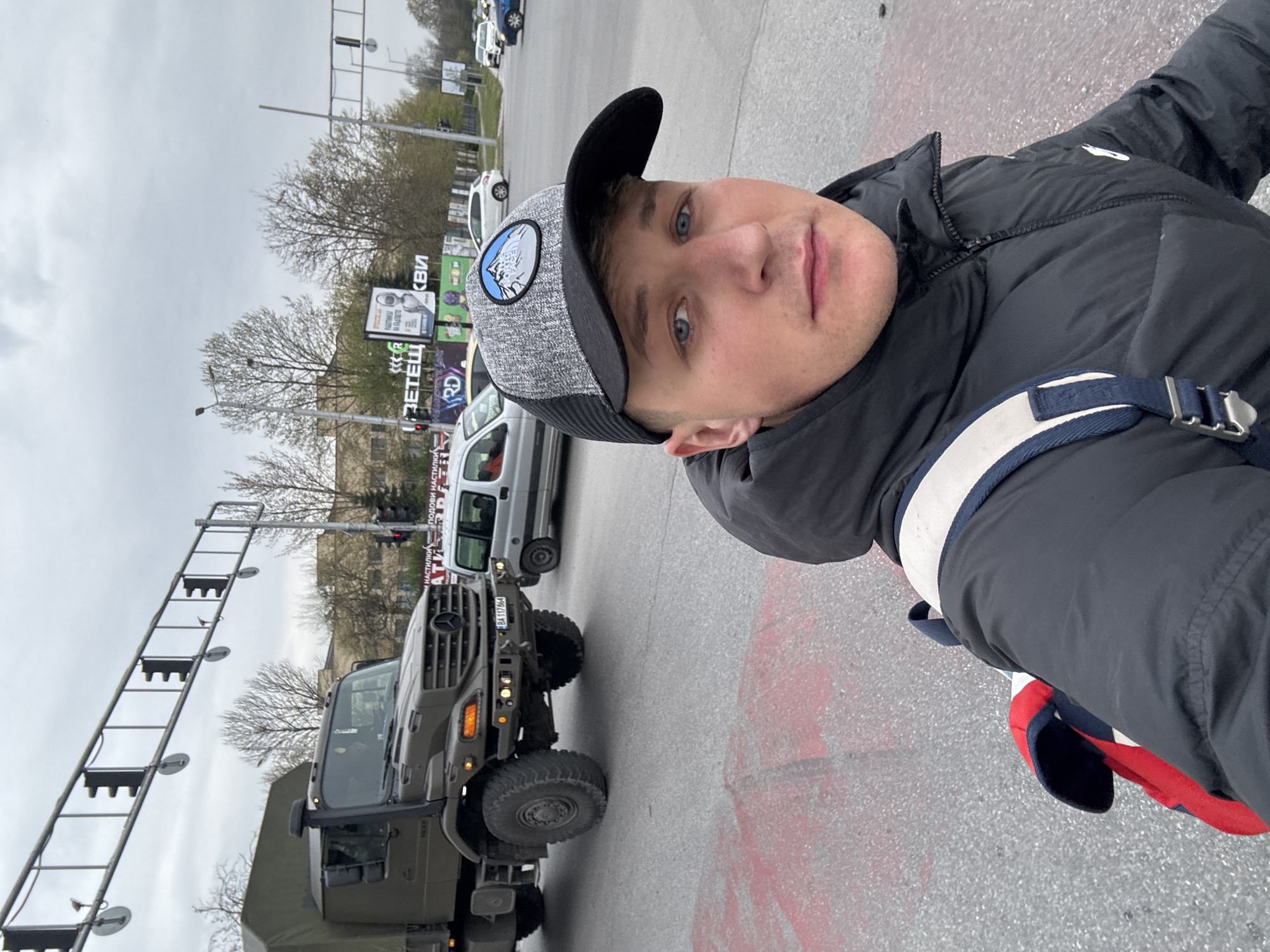 A man is taking a selfie on a street in Plovdiv, Bulgaria. The man is wearing a black jacket, a gray cap with a patch, and a shoulder bag with a blue and white strap. Behind him is a military vehicle, a white van, a billboard, traffic lights, and bare trees. The street is paved with gray asphalt and has some red markings. The sky is overcast.