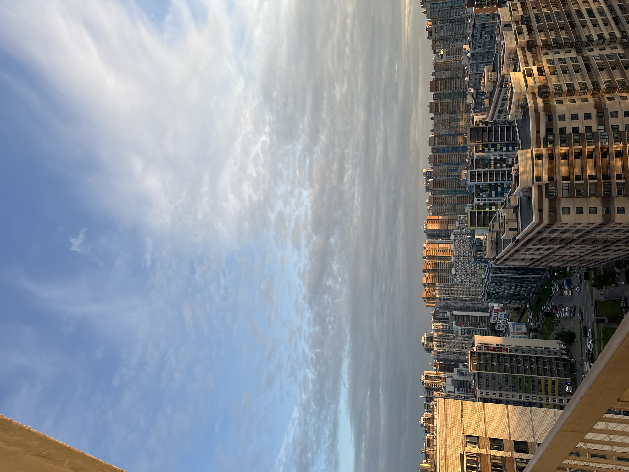 The image shows a view of a densely populated urban area with numerous multi-story apartment buildings. The buildings vary in height and architectural style, with some featuring colorful facades and others presenting more uniform, modern designs. The sky above is partly cloudy with patches of blue visible, suggesting either early morning or late afternoon light. The foreground includes a portion of a balcony railing, indicating the vantage point is from an elevated position. The overall impression is of a large residential district within a city. The location of Feklistovo, Russia, cannot be confirmed from the visual information provided.