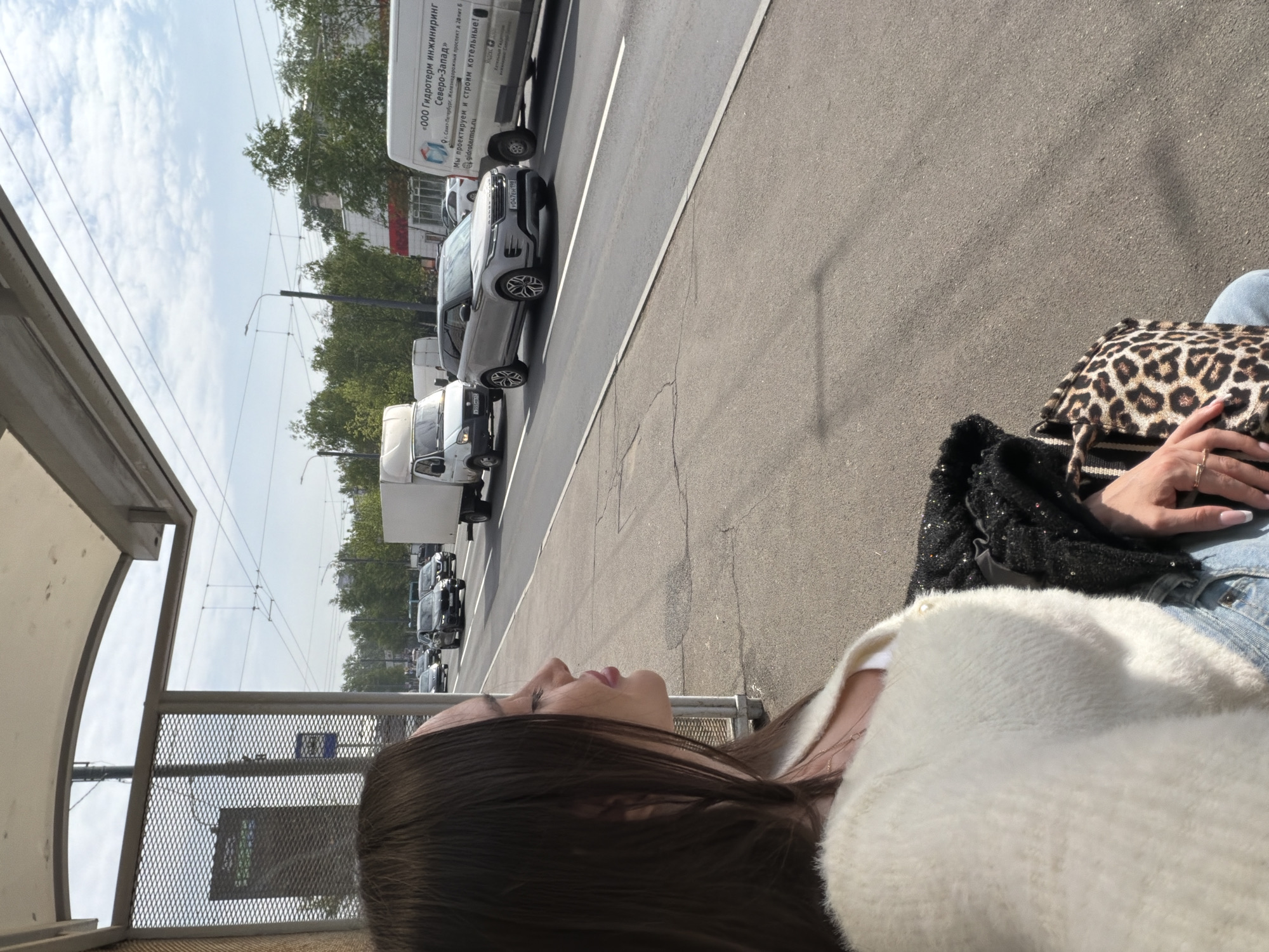 The image shows a woman sitting at a bus stop in Saint Petersburg, Russia. She has dark hair and is wearing a white sweater and blue jeans. She is carrying a leopard-print purse. The bus stop is located on a busy street with cars and trucks passing by. A truck with a sign in Russian, “ООО Гидротерм инжиниринг Северо-Запад”, is visible. The sign references the company’s website, gidrotermsz.ru, and the company’s role in designing and constructing boiler rooms.