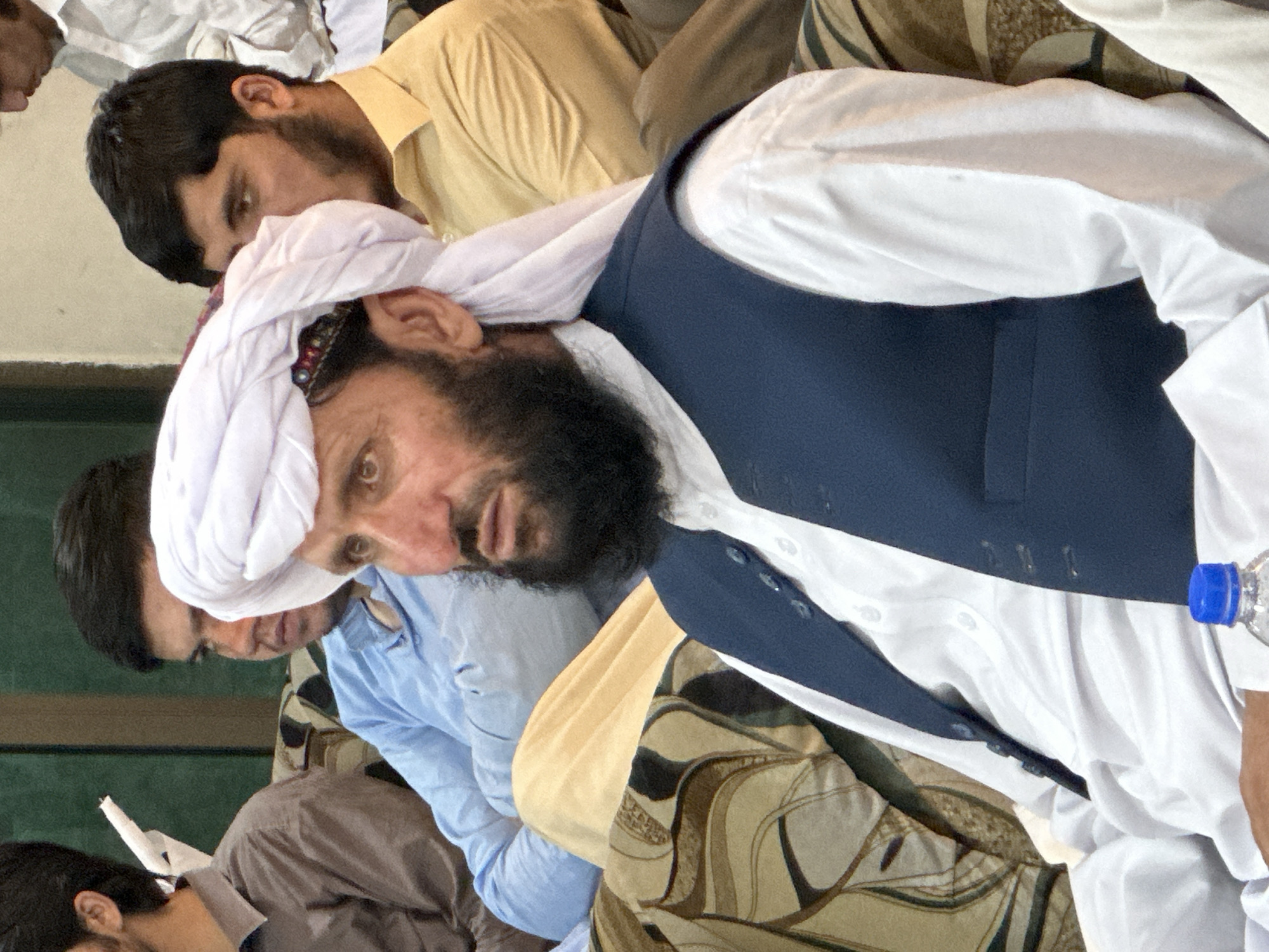 Captured in Quetta City Tehsil, Pakistan, the image presents a gathering of individuals in an indoor setting. Predominantly males are seen, several wearing traditional attire, including turbans and waistcoats over white shirts. One man, central to the frame, wears a white turban and a dark blue waistcoat. The scene includes details of garments and headwear, as well as the facial features of the people present. A bottle of water is also visible on the right. The overall impression is of a meeting or social event taking place in the specified location.