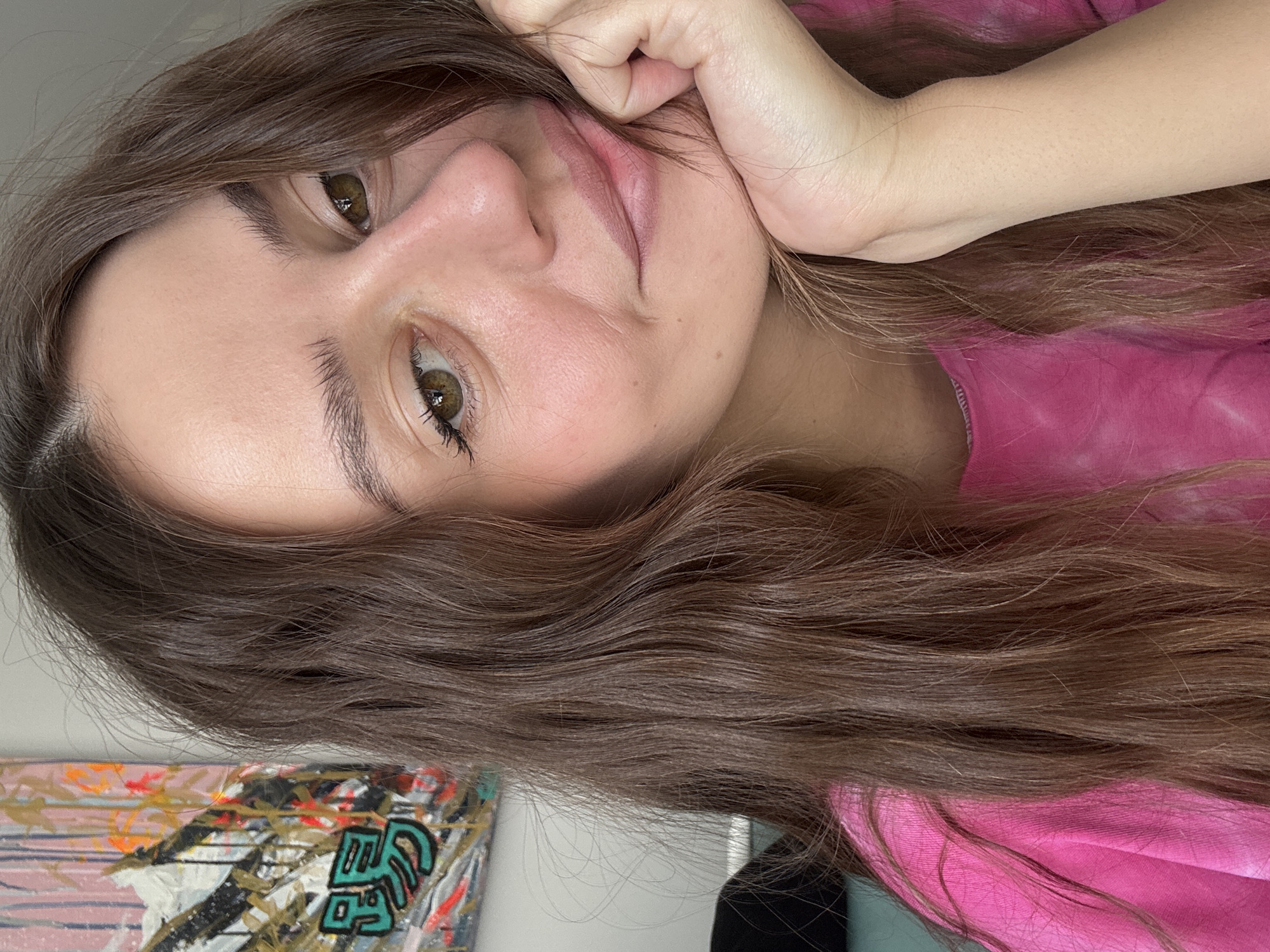 A woman with long brown wavy hair rests her face on her hand, looking at the camera. She wears a pink tie-dye shirt. The background includes a piece of abstract art in the lower left. There is no indication that the image was taken in Krasnoyarsk, Russia.