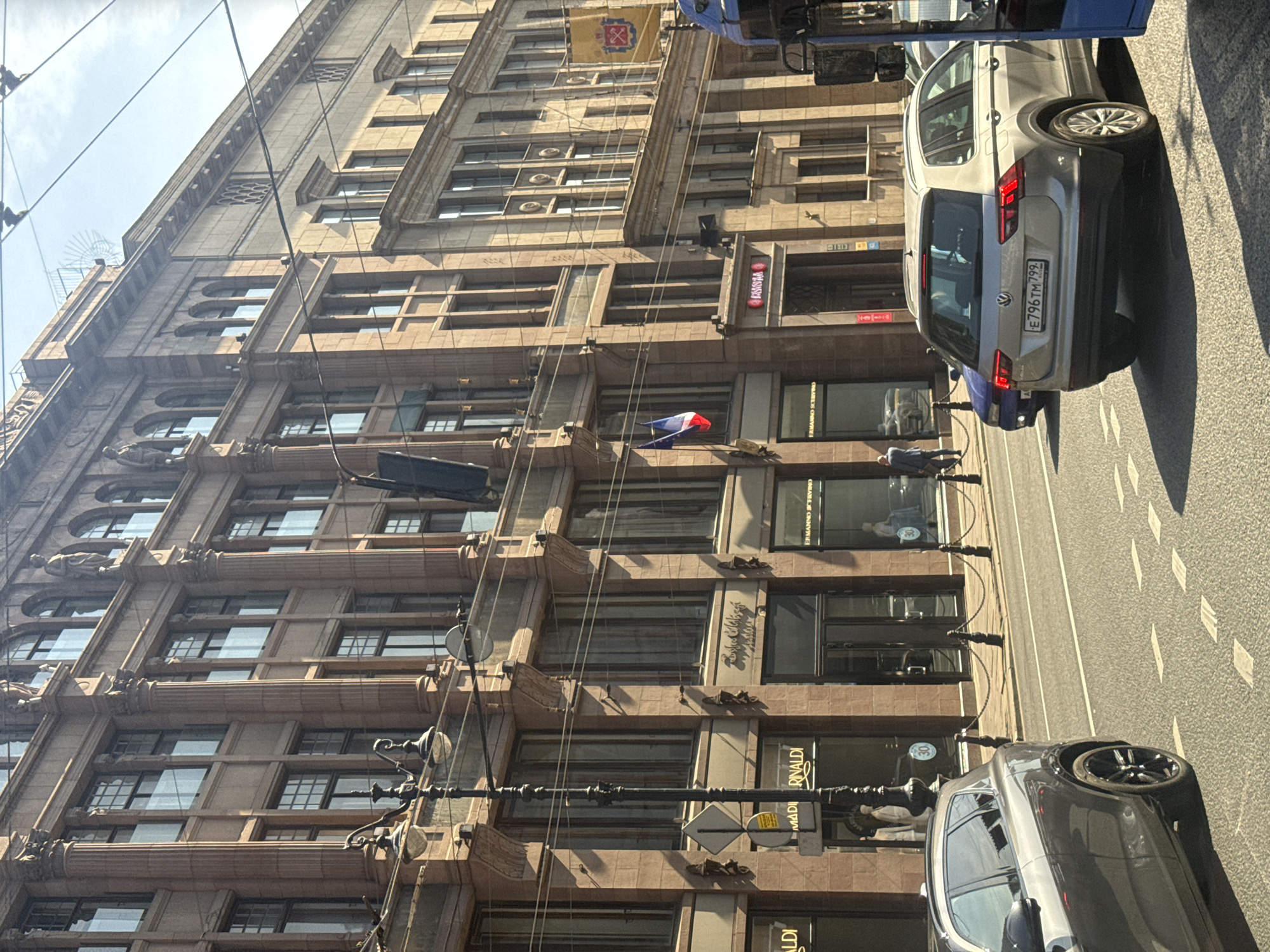 The image shows a street scene in Saint Petersburg, Russia, with a focus on a grand, multi-story building with ornate architecture. The building features large, rectangular windows, decorative columns, and statues on its upper levels. There are several shops at street level, indicated by illuminated signs. Some of these signs are partially visible, showing names like "MADI RINALDI" and "ERMANNO SCERVIN". A lamppost with multiple light fixtures stands on the sidewalk.
The street in front of the building is paved and marked with lane lines. Several cars are present: a dark grey SUV is in the foreground on the left, and a silver Volkswagen Tiguan is in the right lane, facing away from the camera. Another blue car is visible behind the silver SUV. A portion of a blue bus is visible on the far right edge of the image. Two small French flags are hanging from the building's facade. Shadows suggest it is daytime, and the sky is visible above the buildings. The overall impression is of a busy urban environment in a historic district.