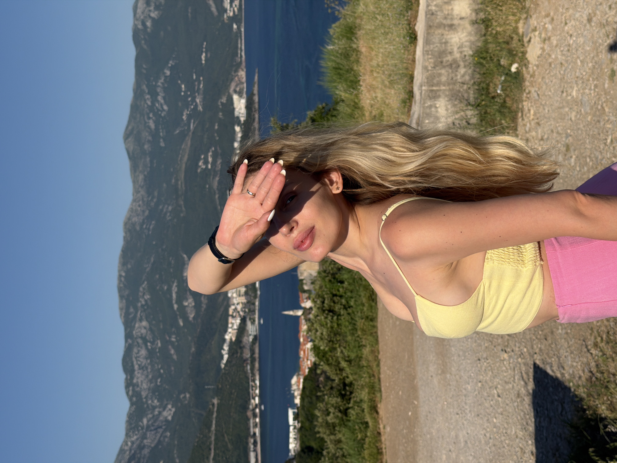 A blonde woman in a yellow top and pink pants shields her eyes with her hand while overlooking a landscape in Budva, Montenegro. The location appears to be an elevated viewpoint near the coast, as mountains, the sea, and a portion of the city are visible in the background. The woman wears a watch on her left wrist and a ring on her left ring finger. The image is oriented with the city in the background on the left and a grassy hillside on the right.