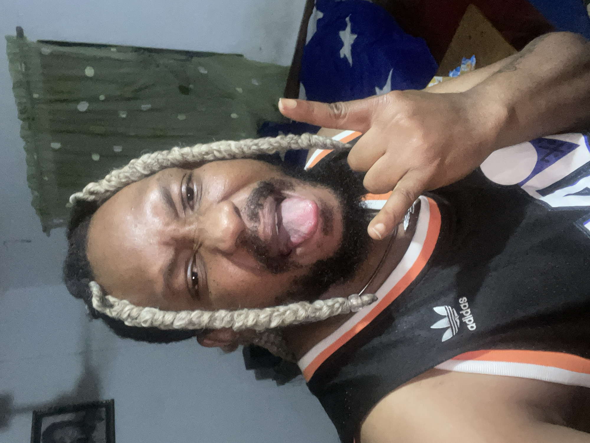 A man with blonde braids, a goatee, and an Adidas jersey is sticking his tongue out and making a rock-and-roll hand gesture. He is indoors, with a green curtain and a framed picture on the wall behind him, and a blue and white star-patterned blanket on a bed in the background. The scene appears to be a bedroom. The provided metadata does not contain information about the specific location within Jos, Nigeria.