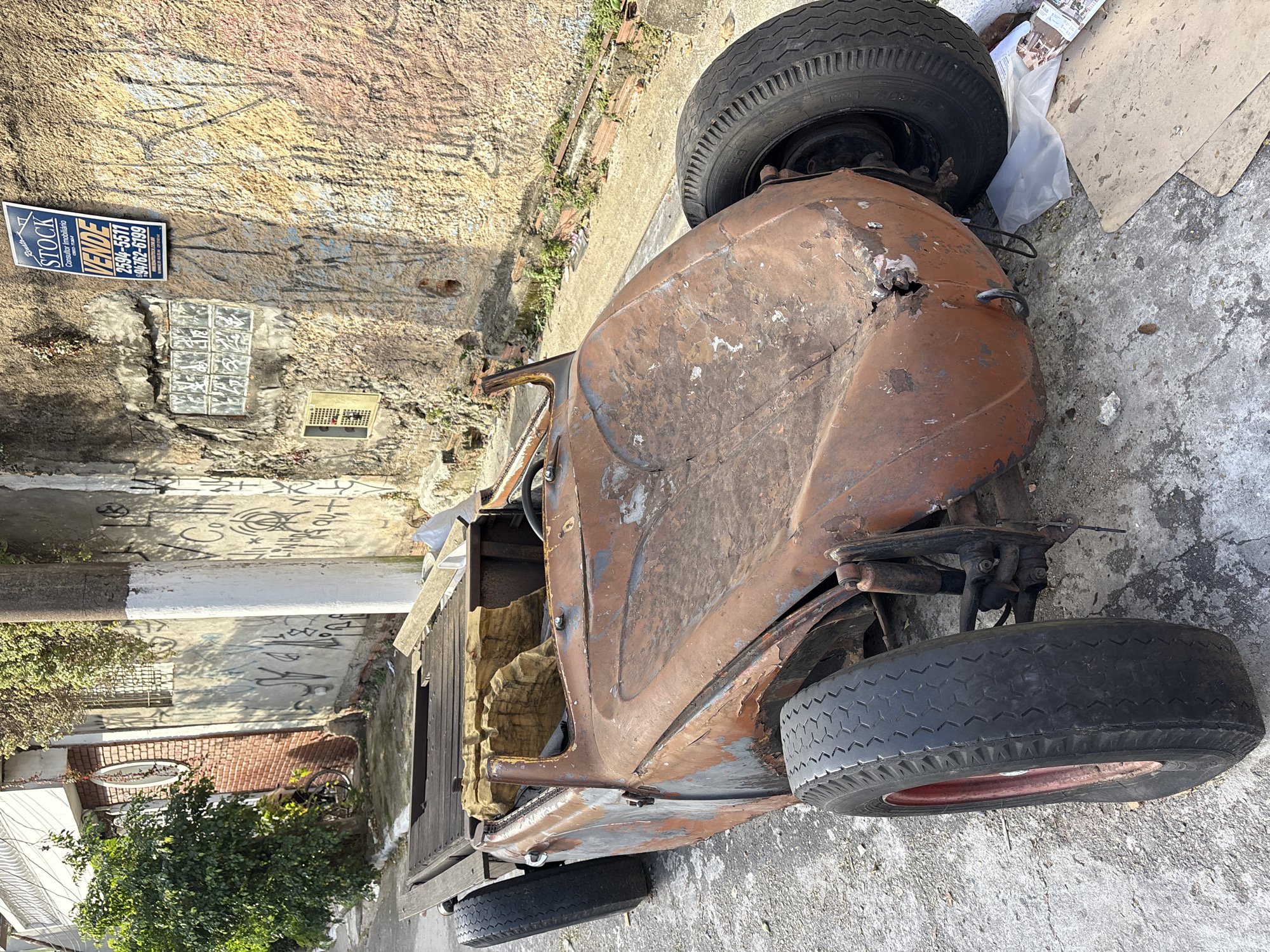 The image shows a rusty, vintage car chassis with a wooden platform on the back, parked on a street in São Paulo, Brazil. The car is a classic model, possibly a Volkswagen Beetle, with its original bodywork significantly deteriorated by rust. The interior is exposed, revealing worn-out yellow upholstery. The car is parked on a paved street with visible cracks and debris. A concrete pole painted with white and black stripes stands to the right of the car. The background features a brick building with a circular window and a wall covered in graffiti, suggesting an urban environment. A "For Sale" sign from "STOCK Consultor Imobiliário" with phone numbers is affixed to the graffiti-covered wall. Green foliage from a tree is visible on the left side of the image. The overall scene depicts a scene of urban decay and neglect.