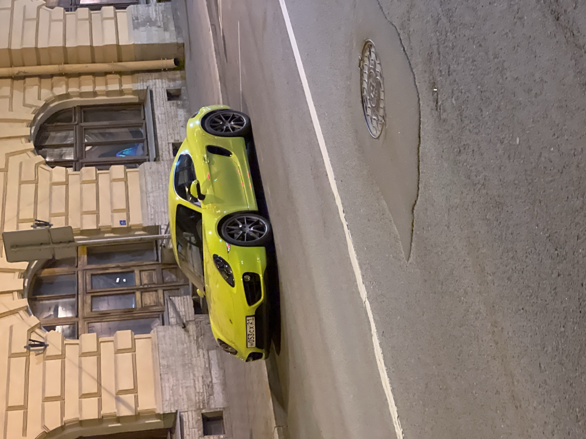 The image shows a yellow Porsche parked on a street in Saint Petersburg, Russia. The license plate number is H053CX 21. The car is parked next to a building with arched windows. There is a manhole cover in the street. The image appears to have been taken at night.