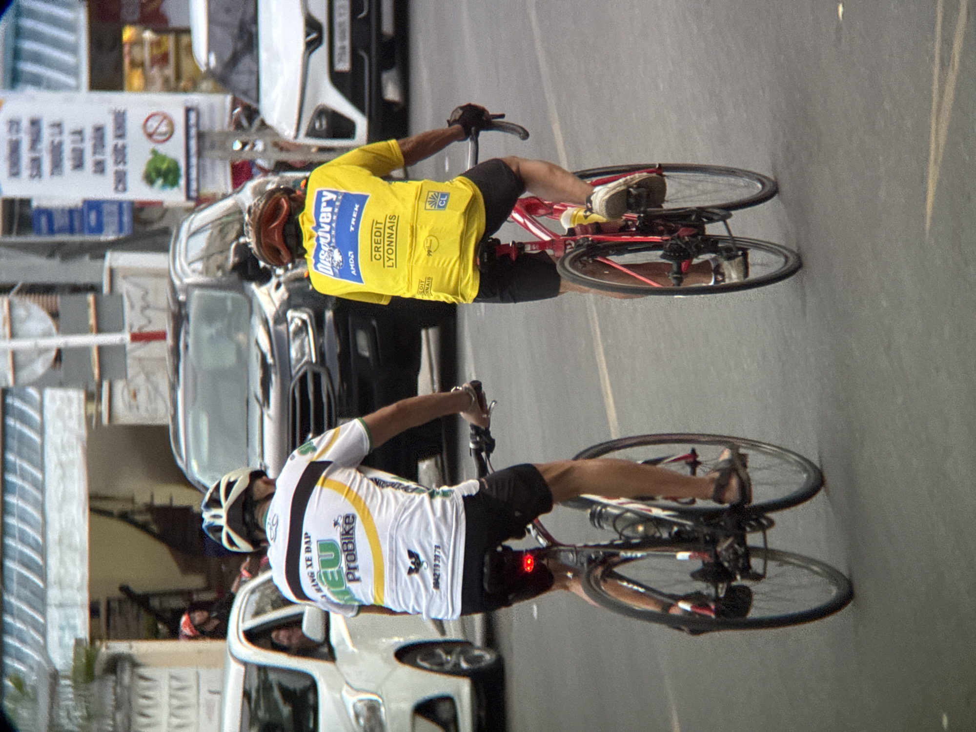 A rear view captures two cyclists riding on an asphalt road. The cyclist on the left wears a white and yellow cycling jersey with "ProBike" logo and a white helmet. The cyclist on the right wears a bright yellow jersey with "Discovery Channel" and "Credit Lyonnais" logos, a red helmet, and black cycling shorts. Both cyclists are in motion, with their legs pedaling. The background shows vehicles, including a white car on the left and a black SUV in the middle, suggesting an urban setting. A blurred building with awnings is visible in the distance. Signs with Vietnamese text are present, including one with an image of a green vegetable and a prohibition symbol. The scene takes place during daytime, with ambient light casting shadows on the road. The overall atmosphere is that of everyday city traffic and recreational cycling.