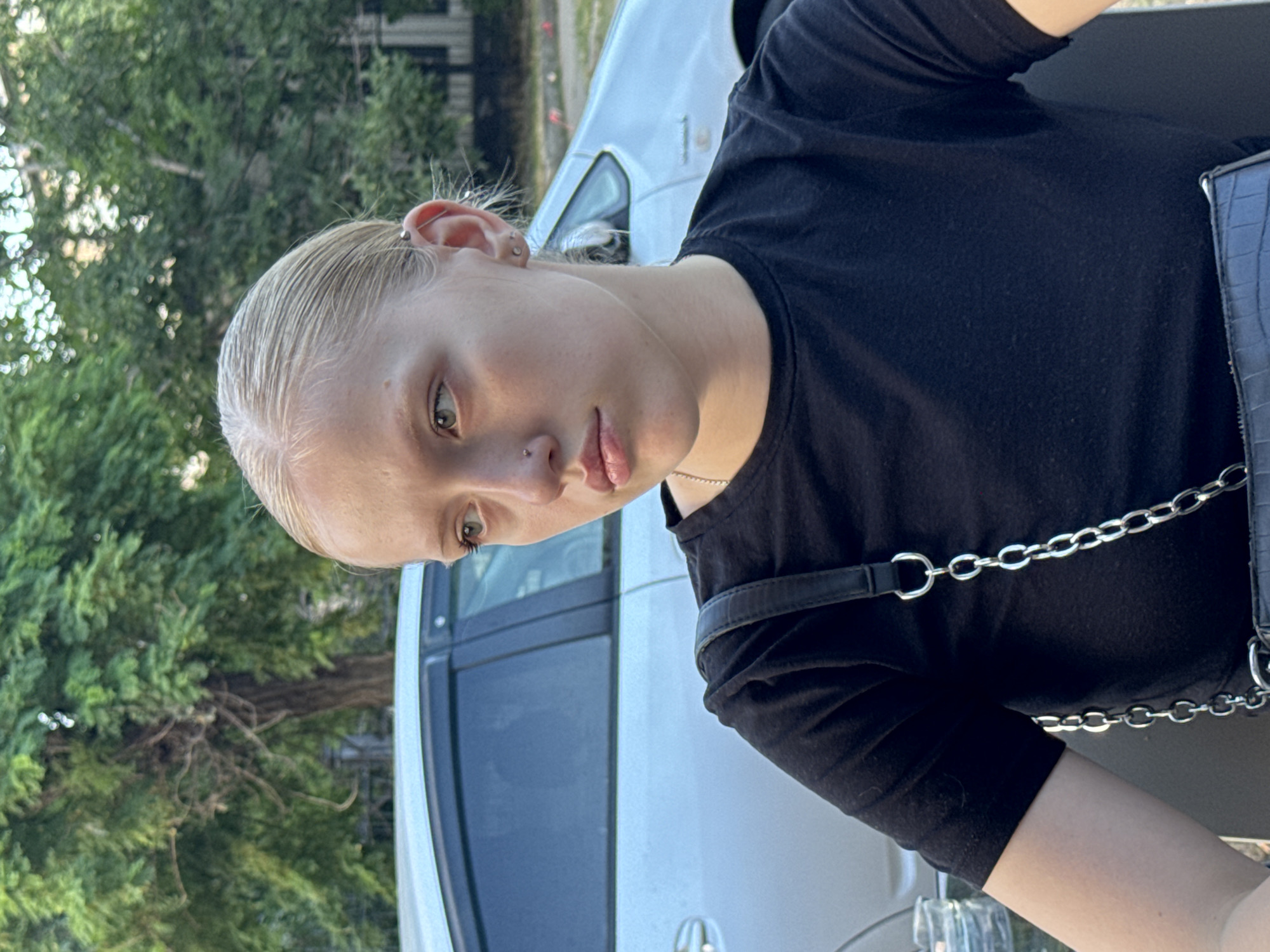 The image shows a close-up, waist-up shot of a young woman with blonde hair pulled back and wearing a black t-shirt. She has a nose ring and earrings. A silver car and green trees are visible in the background. The woman is outdoors, possibly in a park or residential area in Tiraspol, Moldova. Her expression is contemplative as she looks away from the camera. A black handbag with a silver chain strap is visible across her chest.