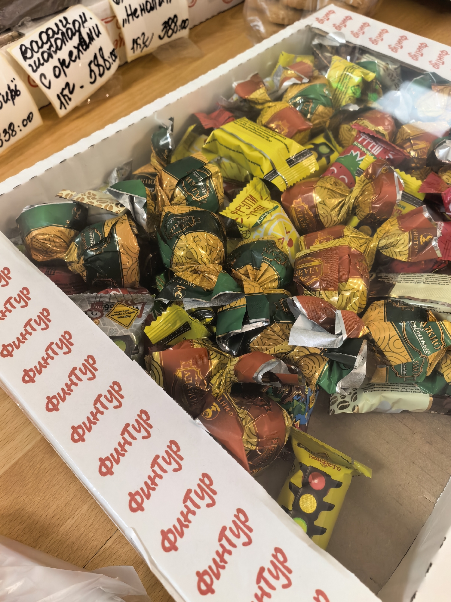 A close-up, eye-level shot captures a cardboard box overflowing with individually wrapped candies. The candies are predominantly gold, green, and red, with some wrappers featuring intricate patterns. A white strip runs along the left side of the box, printed with repeated red text in Russian. In the upper left corner, several price tags are visible, some bearing Russian text and numbers. The background appears to be a wooden surface. The overall scene suggests a retail or market setting, likely a candy stall. Given the context of the city of Pavlovo, Russia, which is known for its confectionery industry, this image likely depicts candies for sale within a shop or market in that city.