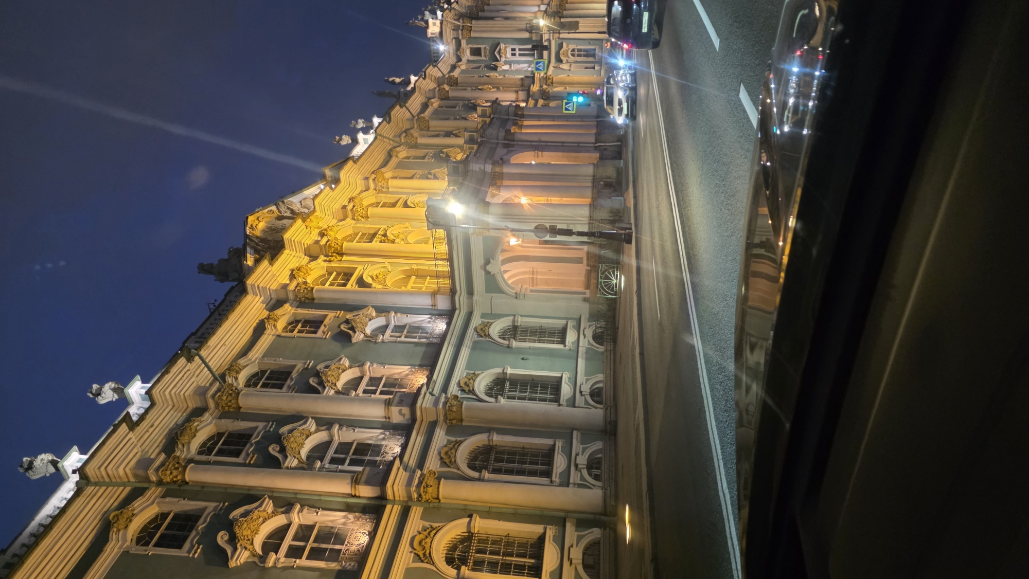 Captured at night in Unknown, Unknown, the photograph features a brightly illuminated, ornate building situated alongside a road bustling with traffic. The building's facade exhibits intricate detailing and is bathed in warm, golden light, contrasting sharply with the darker sky above. The road, marked with distinct white lines, is occupied by several vehicles. The perspective suggests the photo was taken from inside a vehicle, with the side of the car visible in the frame.