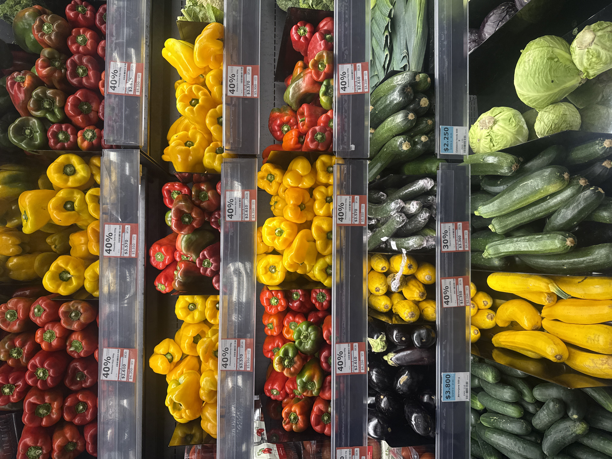The image shows a retail display of various vegetables at a supermarket in Mosquera, Colombia. The display is organized into multiple shelves, each featuring different types of produce. The top shelf is filled with an assortment of red, yellow, and green bell peppers. The subsequent shelves also showcase bell peppers in various colors, alongside eggplants, yellow squash, and zucchini. Some of the produce items have price tags indicating discounts and unit prices. The lighting in the store is bright, illuminating the colorful vegetables. The overall environment suggests a well-stocked grocery store with fresh produce readily available to customers.