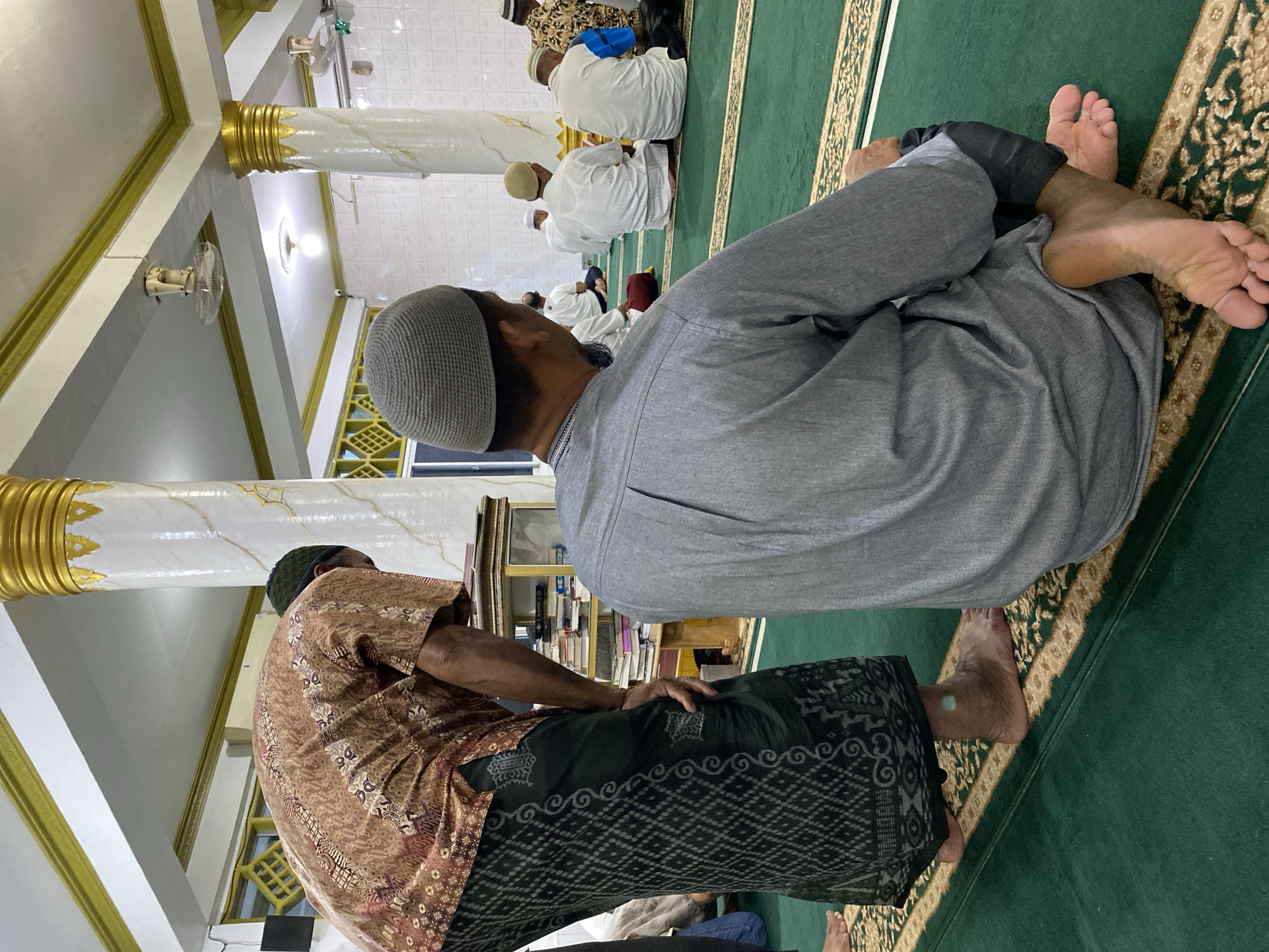 The image shows a gathering of people inside a mosque, likely in Lhokseumawe, Indonesia. Several men are present, some wearing traditional Muslim attire, including skullcaps. They are seated on a green prayer rug, possibly engaged in prayer or a religious activity. The mosque's interior features white pillars with gold accents, indicating a place of worship. Details include the lighting fixtures and the ornate design of the prayer rug. A bookshelf is visible in the background, suggesting a space for religious texts.