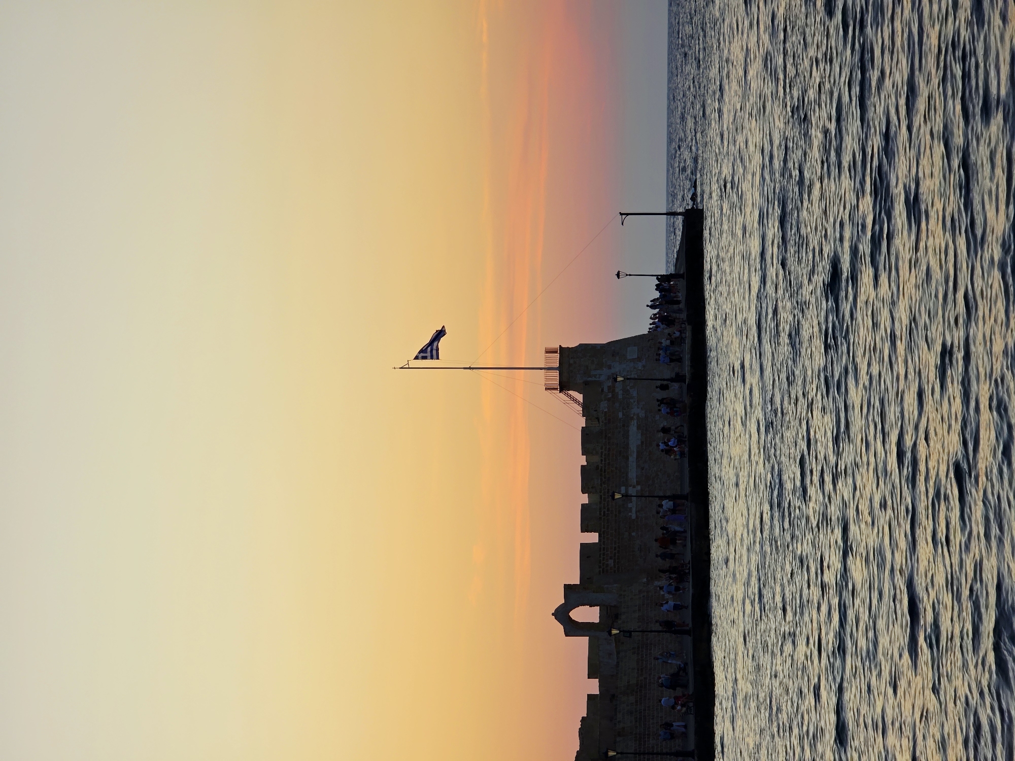 The image captures a sunset view of the Venetian harbor wall and lighthouse in Chania, Greece. The sky displays a gradient of warm colors, transitioning from pale yellow at the top to soft orange and pink hues near the horizon. The sea in the foreground is a deep blue, with gentle ripples reflecting the sky's colors. Silhouetted against the sky and sea are people strolling along the harbor wall, some gathered near a stone structure with an arched opening. A Greek flag is prominently displayed on a flagpole atop a tower-like structure on the right side of the harbor wall. Several lampposts are also visible along the walkway. The overall scene conveys a tranquil evening atmosphere.