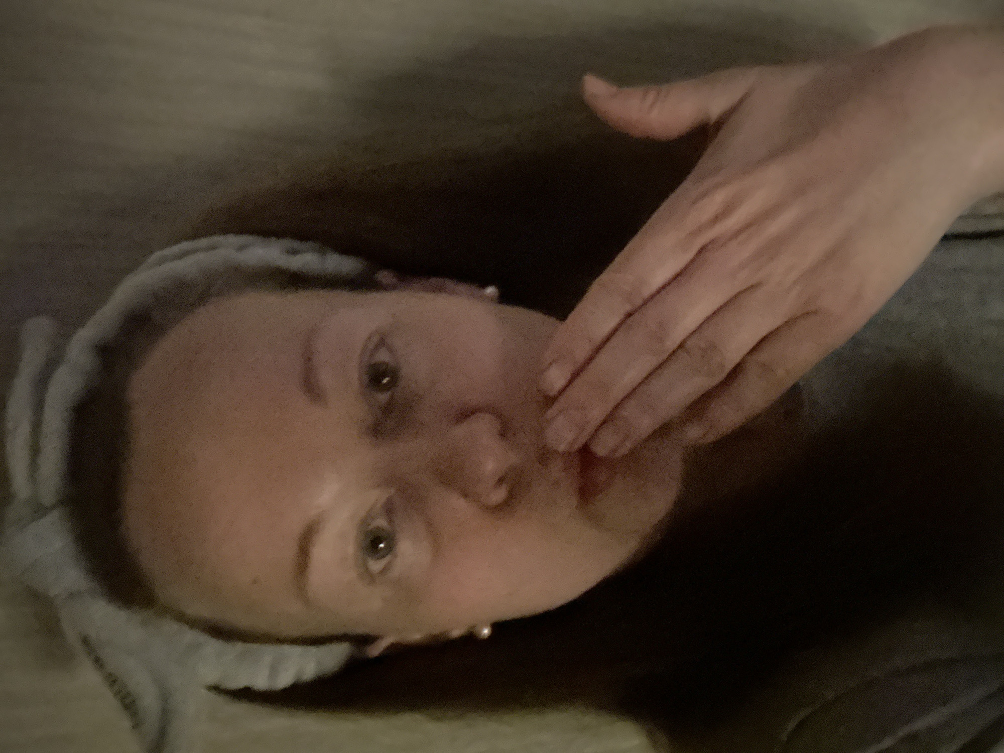 A woman lies on her back, wearing a grey headband and what appears to be a grey shirt. Her hand is positioned over her mouth and cheek. She has fair skin, light eyes, and pearl earrings. The background is unclear, but seems to consist of dark, neutral tones. The image does not contain sufficient information to determine its location in Moscow, Russia, or if it is even located in Moscow, Russia.