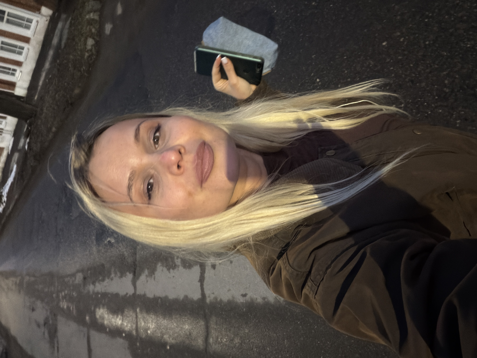 A woman with blonde hair is taking a selfie outdoors, holding a phone in her left hand. The background shows a paved area, likely a sidewalk, with a building visible in the distance. The woman is wearing a dark jacket. A gray object rests next to the phone. The image does not contain information that could identify Sarapul, Russia as the location.