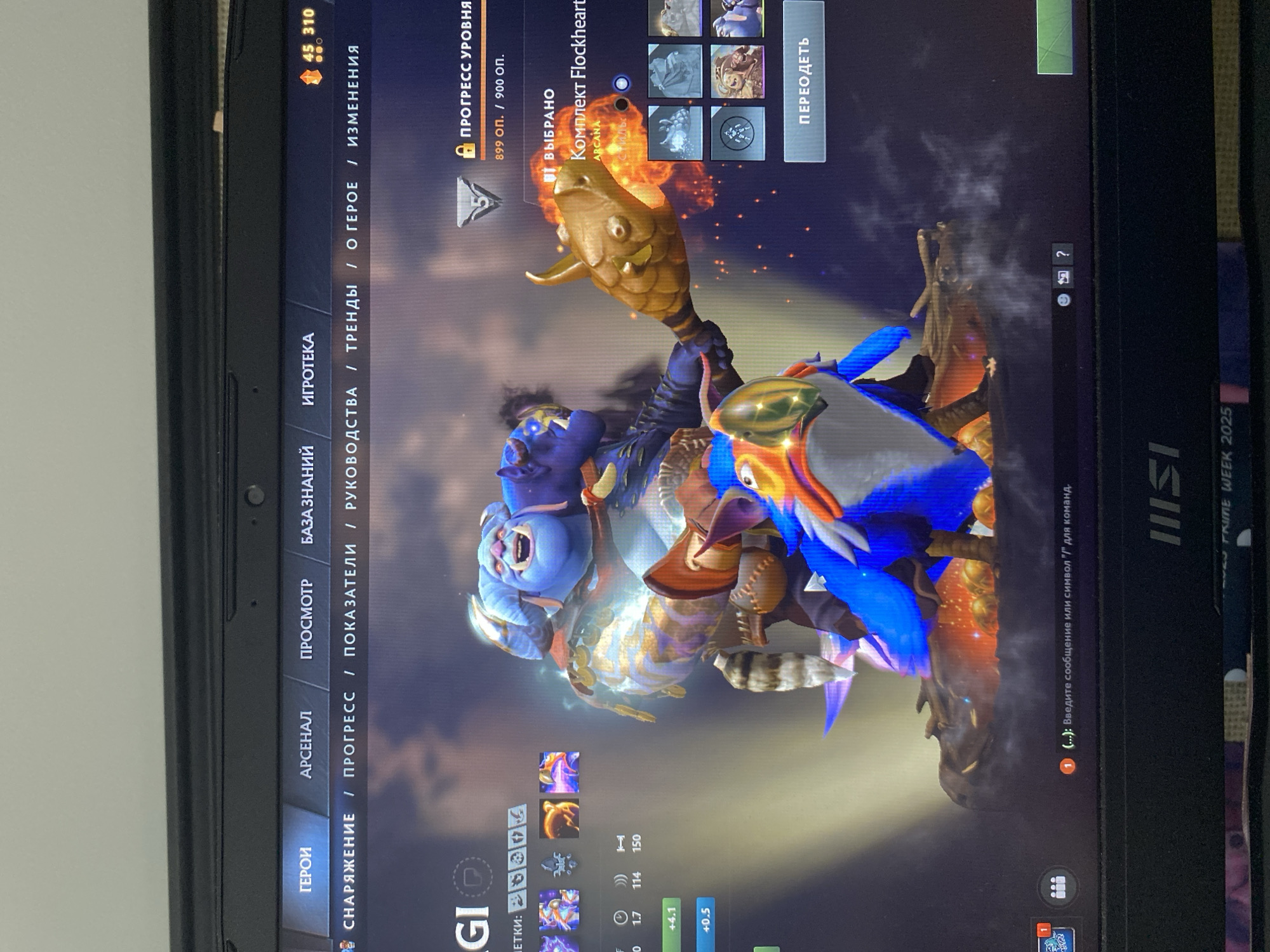 This image is a screenshot of a video game, likely Dota 2, displayed on a laptop screen. The game features a character with blue skin and large ears, adorned with golden armor and what appears to be a fish-like weapon. The character is surrounded by fiery effects. In the foreground, there are several icons representing in-game items or abilities.
The laptop screen shows a user interface with various game-related menus and information. To the left of the character, there are tabs labeled "ГЕРОИ" (Heroes), "АРСЕНАЛ" (Arsenal), "ПРОСМОТР" (View), "БАЗА ЗНАНИЙ" (Knowledge Base), "ИГРОТЕКА" (Game Library), and other navigation elements. At the top, text in Russian indicates "ПРОГРЕСС УРОВНЯ" (Level Progress) with a numerical value of "899 оп. / 900 оп.". Below this, it states "ВЫБРАНО" (Selected) and mentions "Комплект Flockheart" (Flockheart Set) with "ARCANA" highlighted. A button labeled "ПЕРЕОДЕТЬ" (Redress/Equip) is also visible.
In the lower part of the screen, there are further item slots and stat displays. The bottom right corner shows the MSI logo and "PRIME WEEK 2025", suggesting the laptop's brand and possibly a promotional event. The presence of Russian text implies the game is being played with a Russian language setting. The overall scene is a digital interface within a video game, with no indication of a specific time of day, weather, or real-world location beyond the laptop itself. The character's expression is animated and suggests action or display within the game.