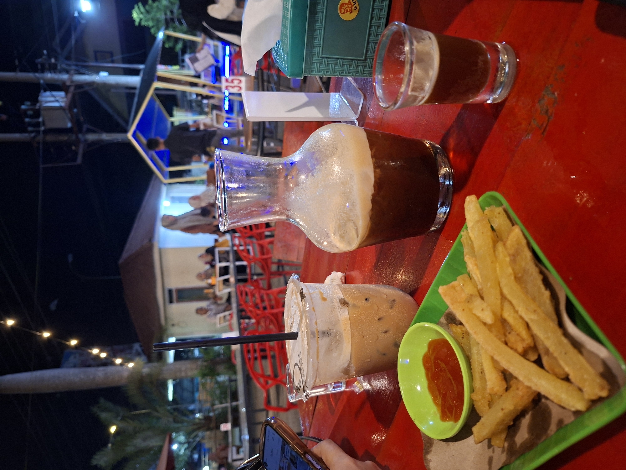 On a red table in Lhokseumawe, Indonesia, are food and drinks. A glass pitcher filled with a dark liquid with foam sits in the middle of the table. To its right is a shot glass with a similar dark liquid. A green tissue box sits behind it. To the left is a plastic cup with a brown beverage and a black straw. Next to it is a green triangular plate of french fries and a small dish of red sauce. A hand holding a phone is visible in the lower left corner. In the background, red chairs and people are present under dimly lit awnings. String lights hang above. A blue sign with the number 35 is visible above.