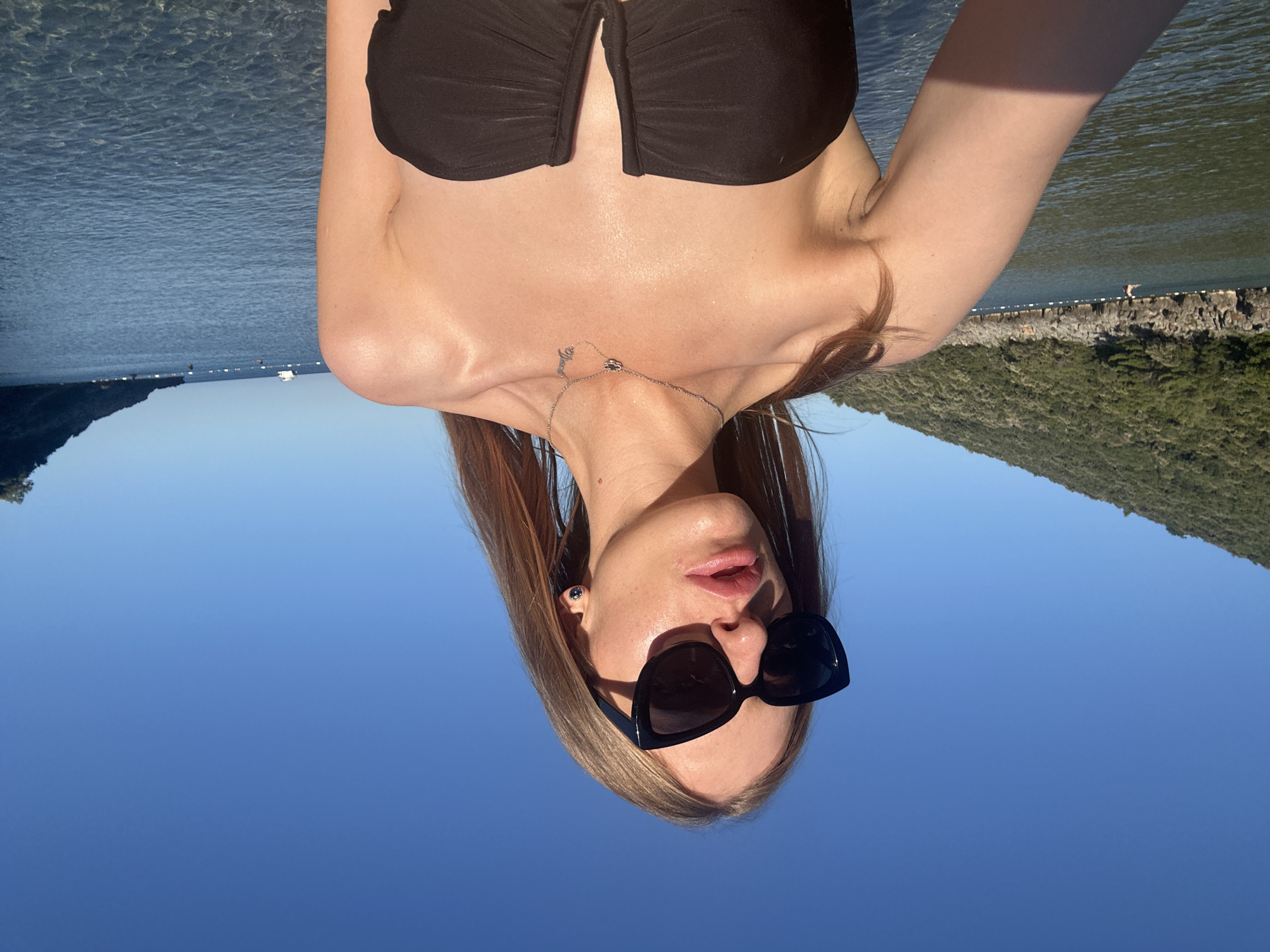 A young woman with long, straight light brown hair, wearing black cat-eye sunglasses and a black strapless bikini top, is depicted in a selfie taken in the clear, calm waters of Lastva Grbaljska, Montenegro. Her lips are slightly parted, revealing a glossy pink tint, and she wears a small, dark stud earring in her left ear. A slender silver necklace, featuring a small charm and a word pendant, rests on her collarbone. The water is translucent, with the seabed visible in the foreground, reflecting the bright blue sky above. To the left, a verdant, tree-covered hillside with a rugged rocky shoreline extends into the bay. A single swimmer is visible near the shore, within an area marked by white buoy lines. To the right, another distant, forested landmass frames the water, with several small boats anchored further out, including a distinct white vessel. The scene is brightly illuminated by natural sunlight, casting highlights on the woman's skin and the water's surface.