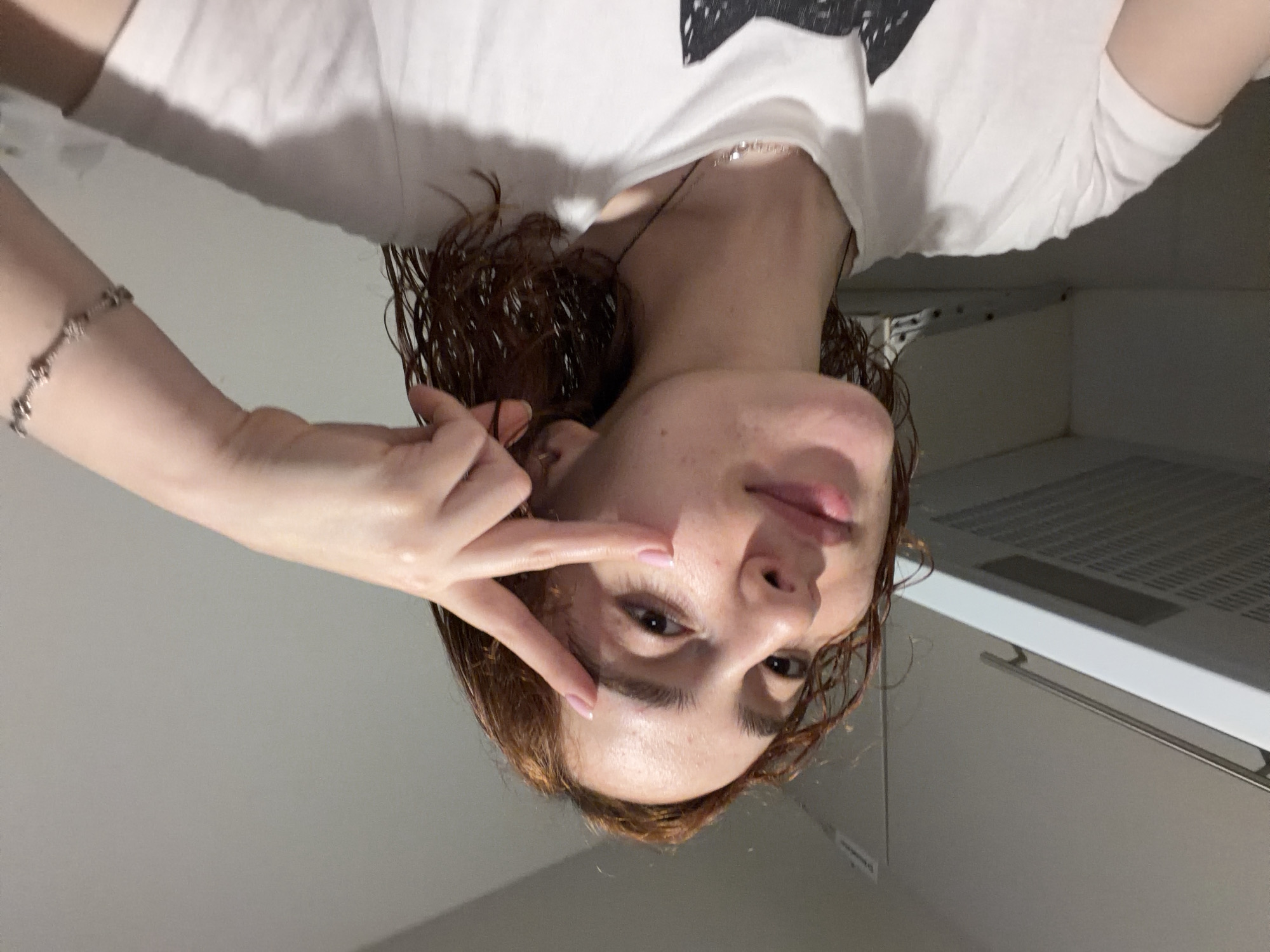 A young woman with reddish-brown, possibly wet or damp, medium-length hair is captured in a close-up, rotated portrait. She appears to be looking directly into the camera. Her right hand is raised, with her index and middle fingers extended in a V-sign gesture near her right eye. She has a neutral to slightly smiling expression, with some freckles or minor blemishes visible on her skin.
She is wearing a white short-sleeved t-shirt, part of which features a dark graphic design on the upper shoulder area. A delicate silver-colored chain necklace is visible around her neck. On her left wrist, she wears a silver-toned bracelet with multiple spherical or circular links. Her fingernails have a light pink or nude polish.
The background suggests an indoor setting, possibly a modern kitchen or utility area. A light-colored wall dominates the background. To the right, a white range hood or ventilation unit with a metallic grate and a dark horizontal panel is visible. Below it, a metallic handle or rail extends across what appears to be a cabinet or appliance. The lighting is even, suggesting artificial illumination. The image is taken in a way that the subject appears upside down, indicating a non-standard camera orientation or pose. The general setting is simple and clean, located in Strasbourg, France. No discernible text is visible in the image.